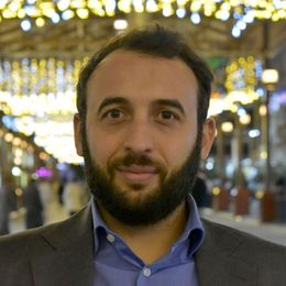 Un uomo con la barba sorride alla telecamera, indossando una camicia blu e un blazer grigio, sullo sfondo una strada illuminata.