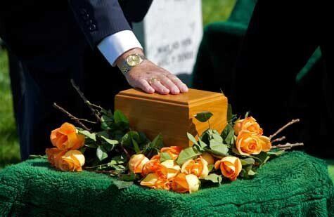 A person’s hand rests gently on a wooden cremation urn surrounded by orange roses on a green cloth at a cemetery.