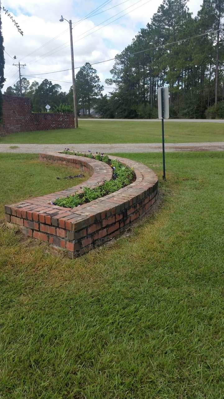 A curved red brick planter filled with small green plants, situated on a green lawn with a street sign in the background.