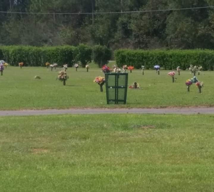 A green trash bin sits in the middle of a sunlit cemetery lawn dotted with flower arrangements and grave markers.