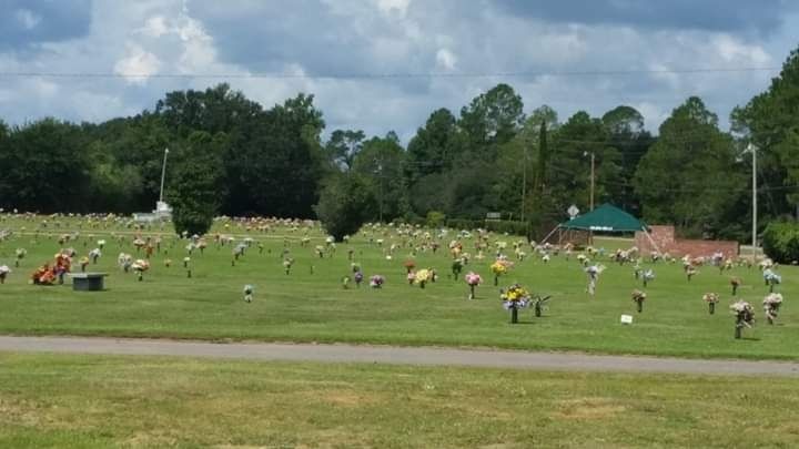 A wide, grassy cemetery landscape with numerous flat grave markers and scattered flower arrangements under a cloudy sky.