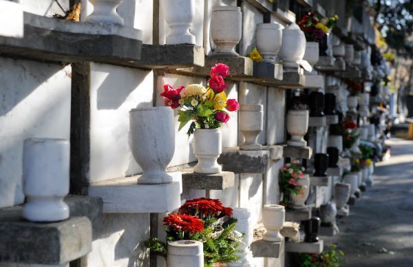 A cemetery columbarium wall with white marble niches and stone vases holding colorful floral arrangements.
