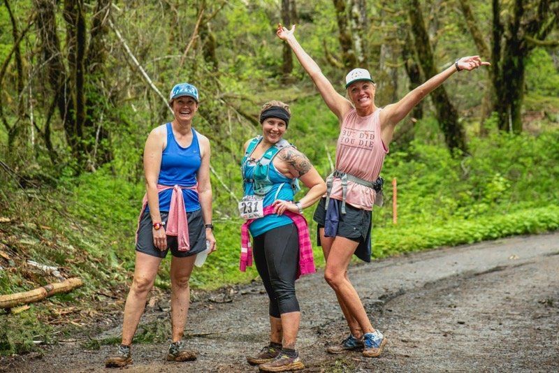 Three women are standing next to each other on a dirt road in the woods.