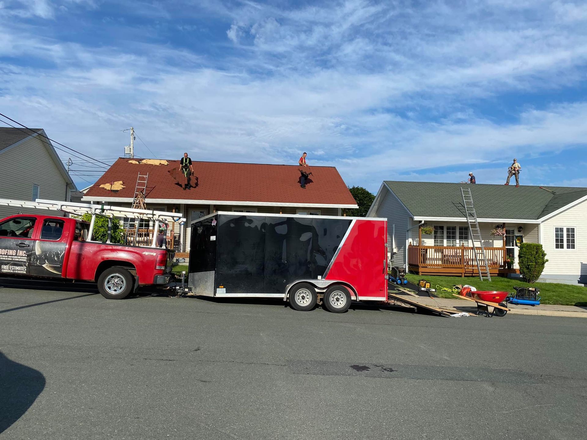 Roofing crew working on houses on a sunny day. Red truck and black trailer parked in front.