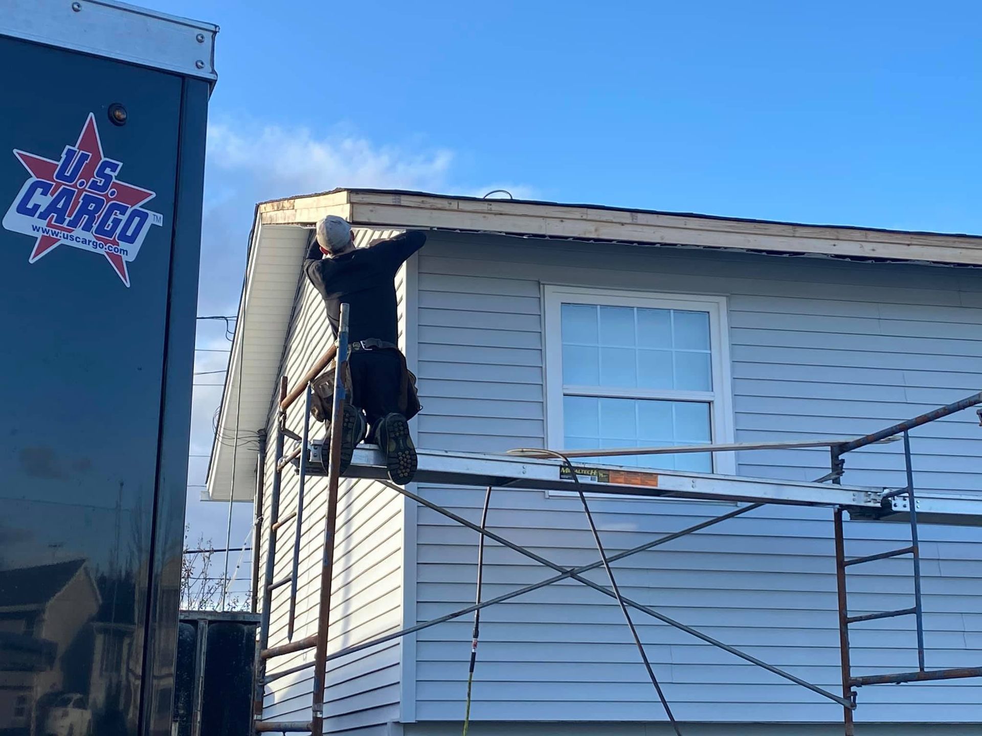 Man on scaffolding repairs a house's roof. Blue sky, light gray siding, and a U.S. Cargo truck are also visible.