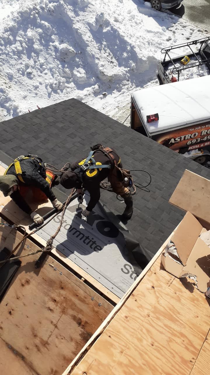 Two roofers in harnesses work on a rooftop, securing tar paper. Snow is present.