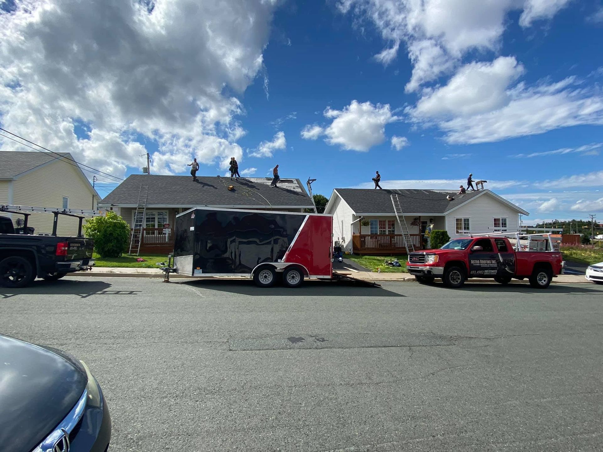 Roofers working on the rooftops of houses on a sunny day, with a trailer, truck, and vehicles parked on the street.
