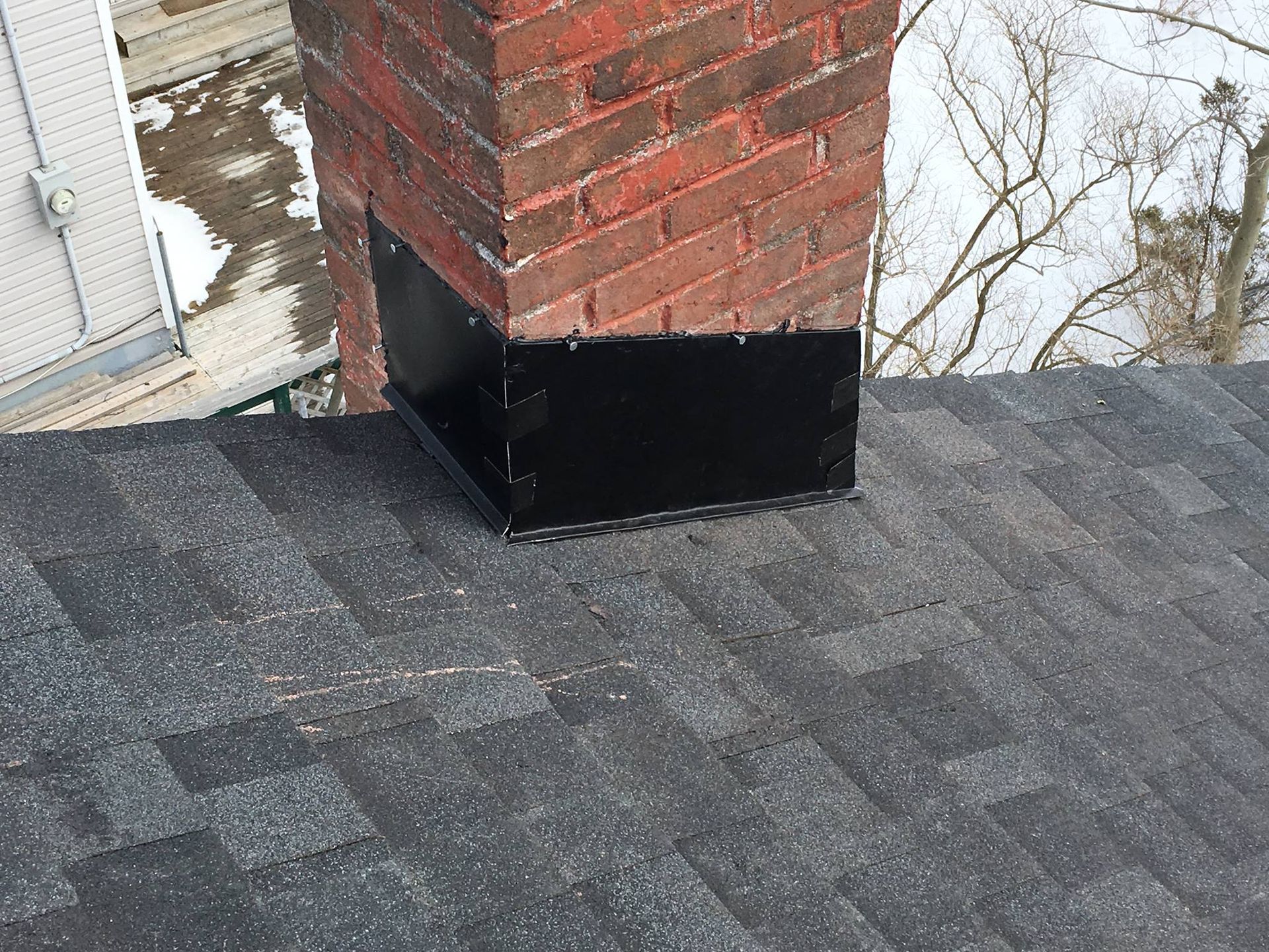 A brick chimney with a black metal flashing sits on a dark gray shingled roof against a snowy winter background.