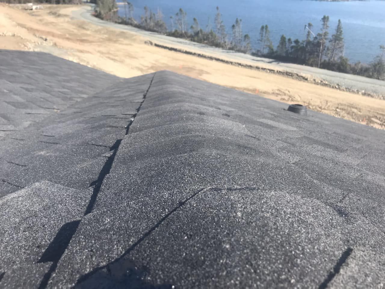 Close-up view of a dark gray shingled roof with a ridge line, with a body of water and land in the background.