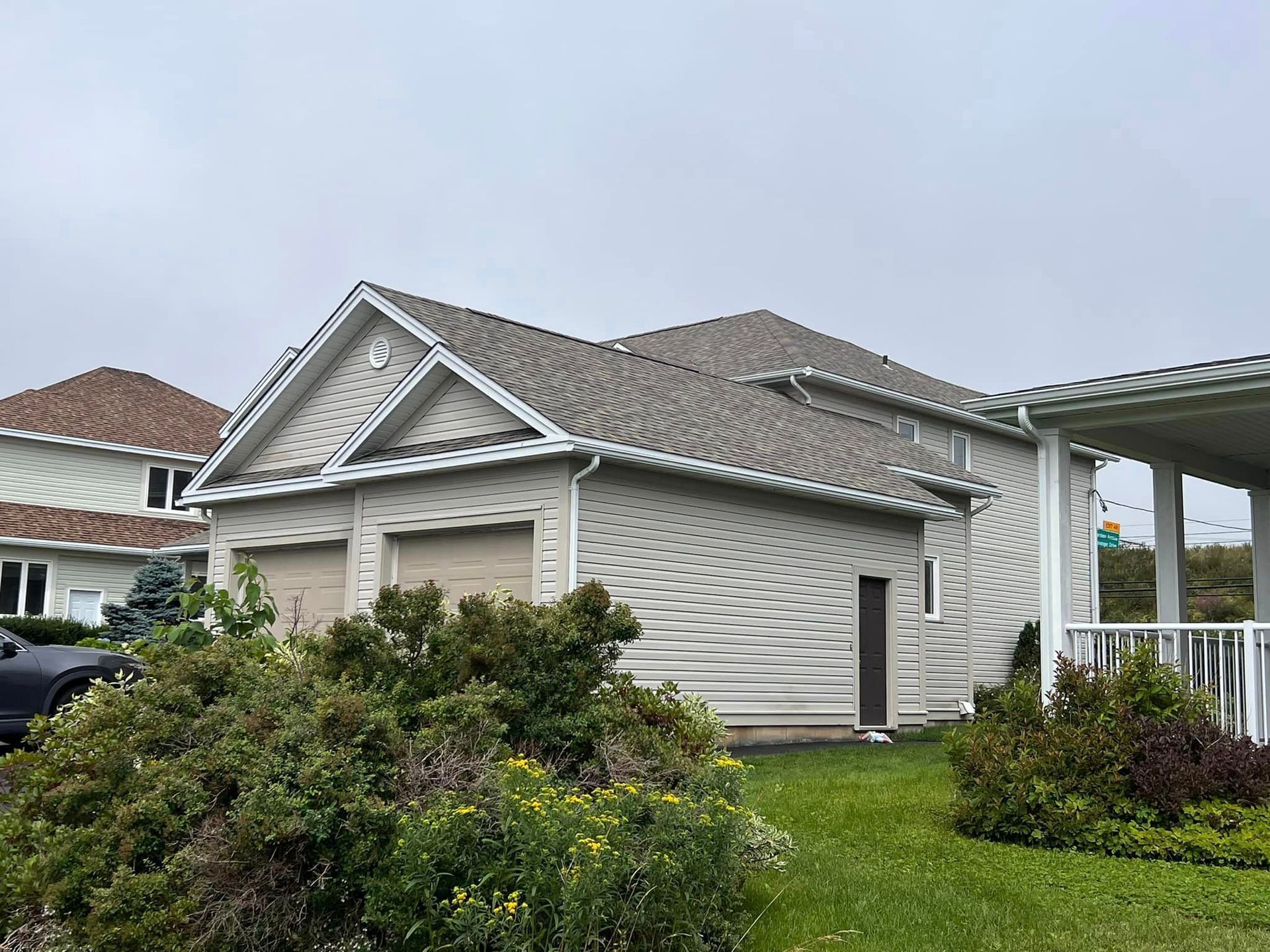 A two-car garage with light siding and a brown roof sits beside a house with a white porch. Overcast sky.