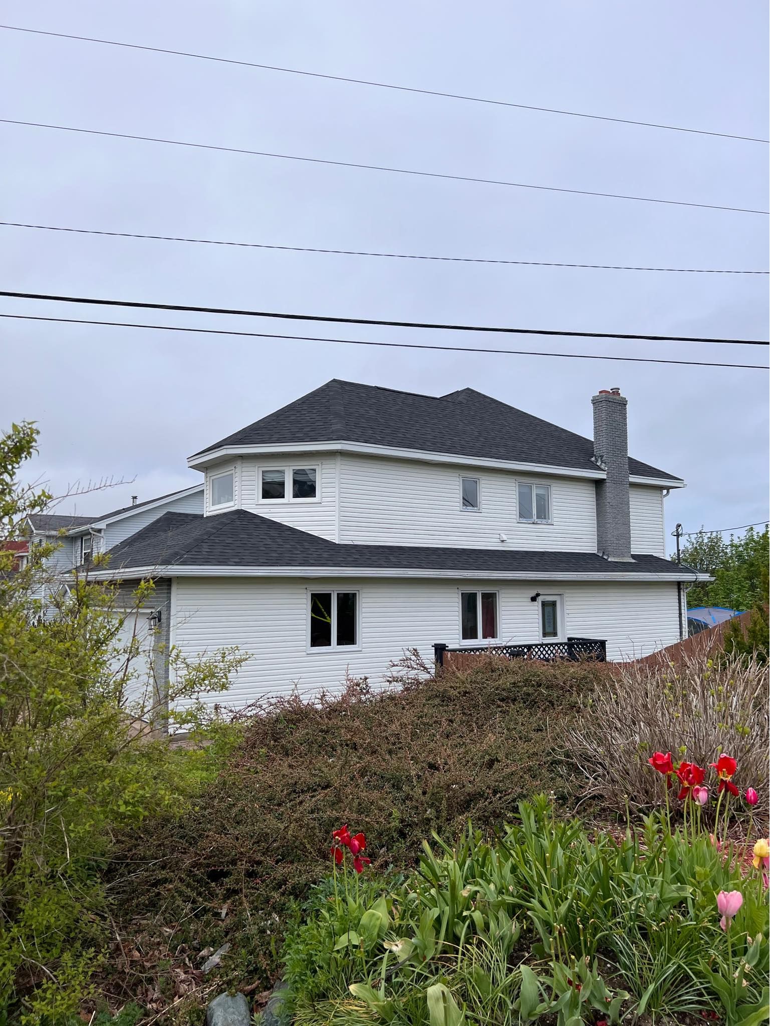 White house with a black roof and tall chimney, seen from a garden on an overcast day. Power lines above.