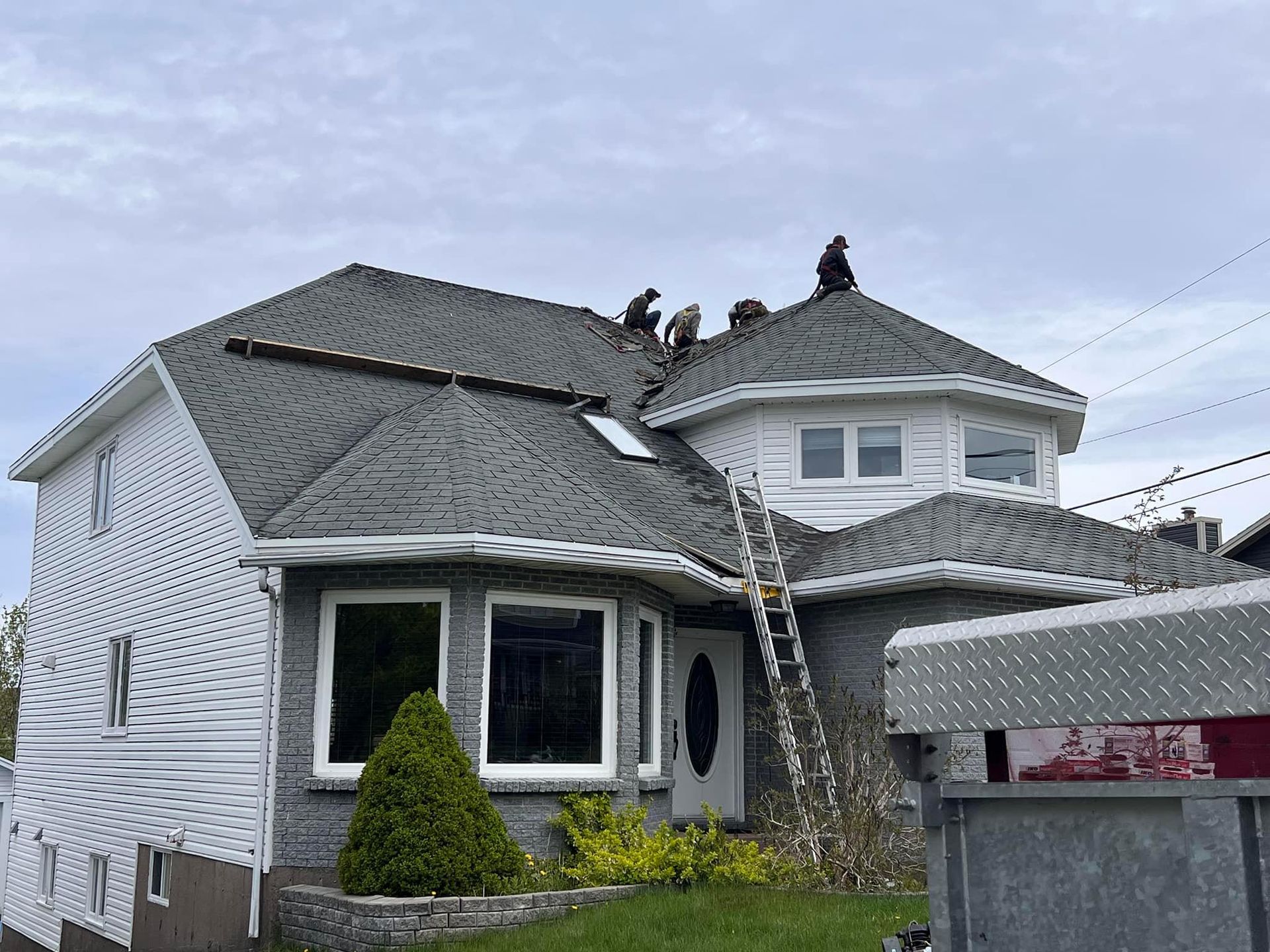 Workers install roofing on a two-story house with gray shingles and white siding. A ladder leans against the front.