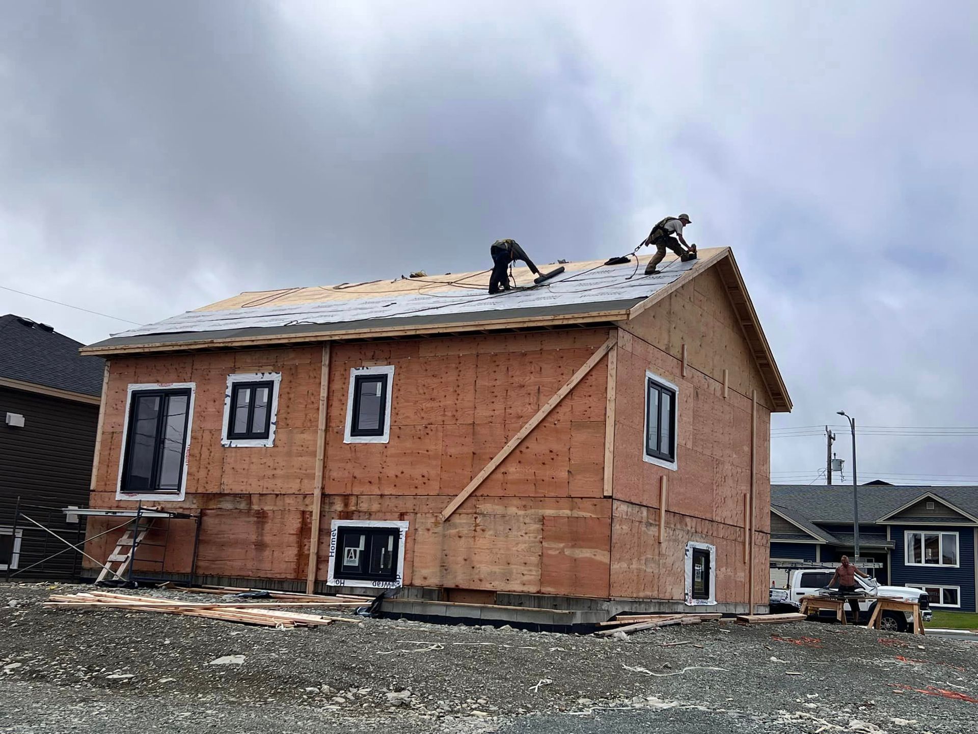 Construction workers installing roofing on a two-story house under a cloudy sky. The house is framed with plywood.
