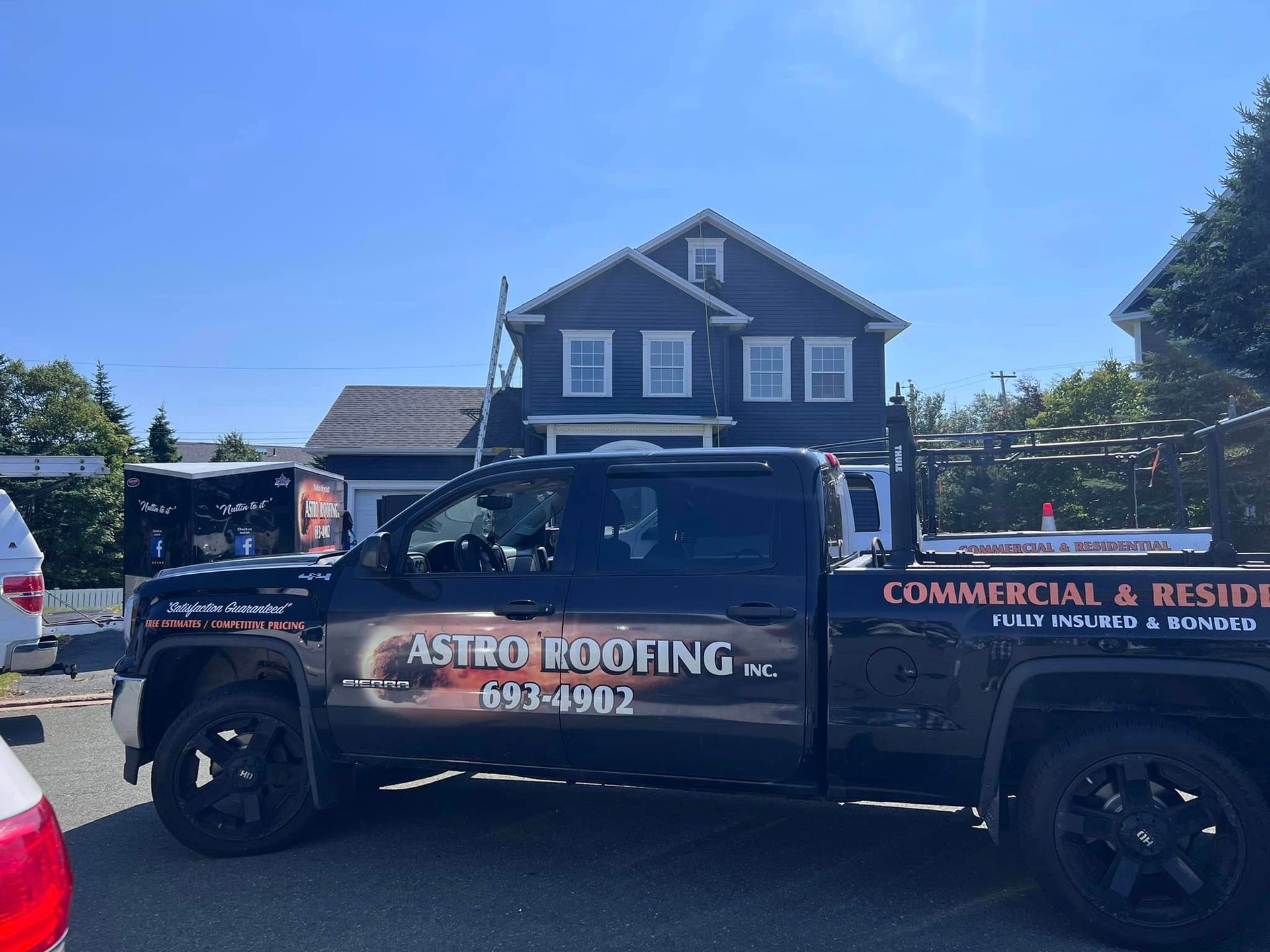 A dark Astro Roofing truck parked in front of a two-story house on a sunny day. The truck has equipment on the back.