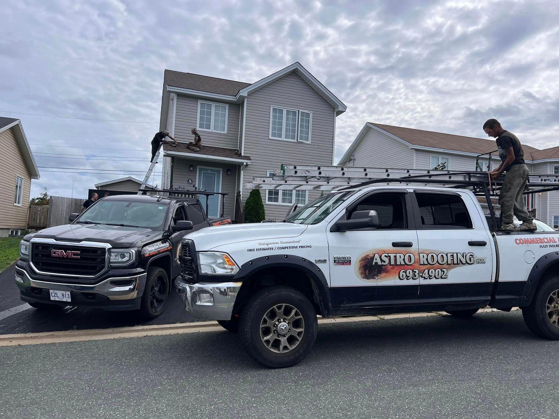 Two work trucks parked in front of a two-story house; one man working on the roof.