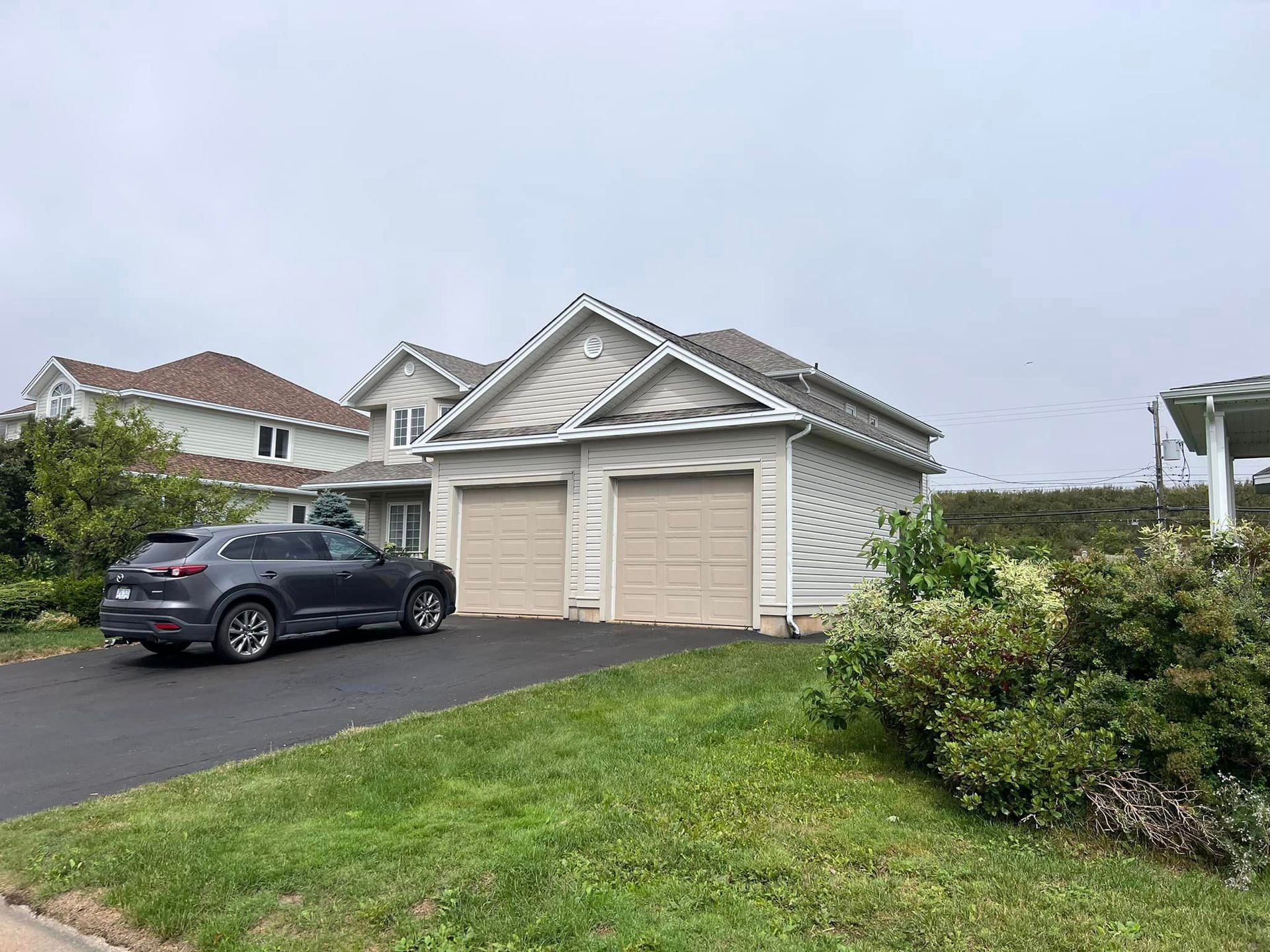 A two-car garage with beige doors sits in front of a gray SUV. A two-story house is visible behind the garage. Overcast day.