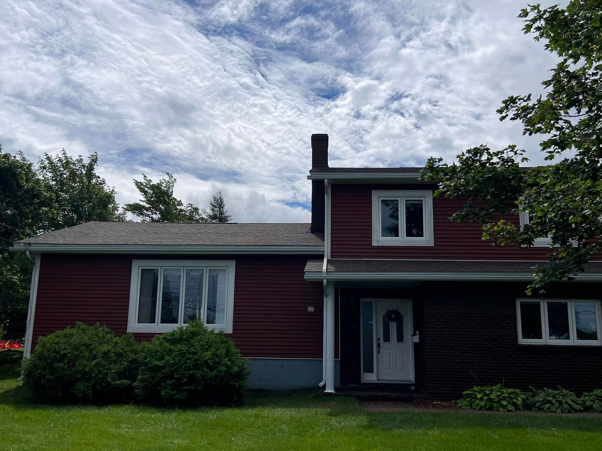 Two-story house with dark red siding, white windows, and a dark door. Green lawn and partly cloudy sky.