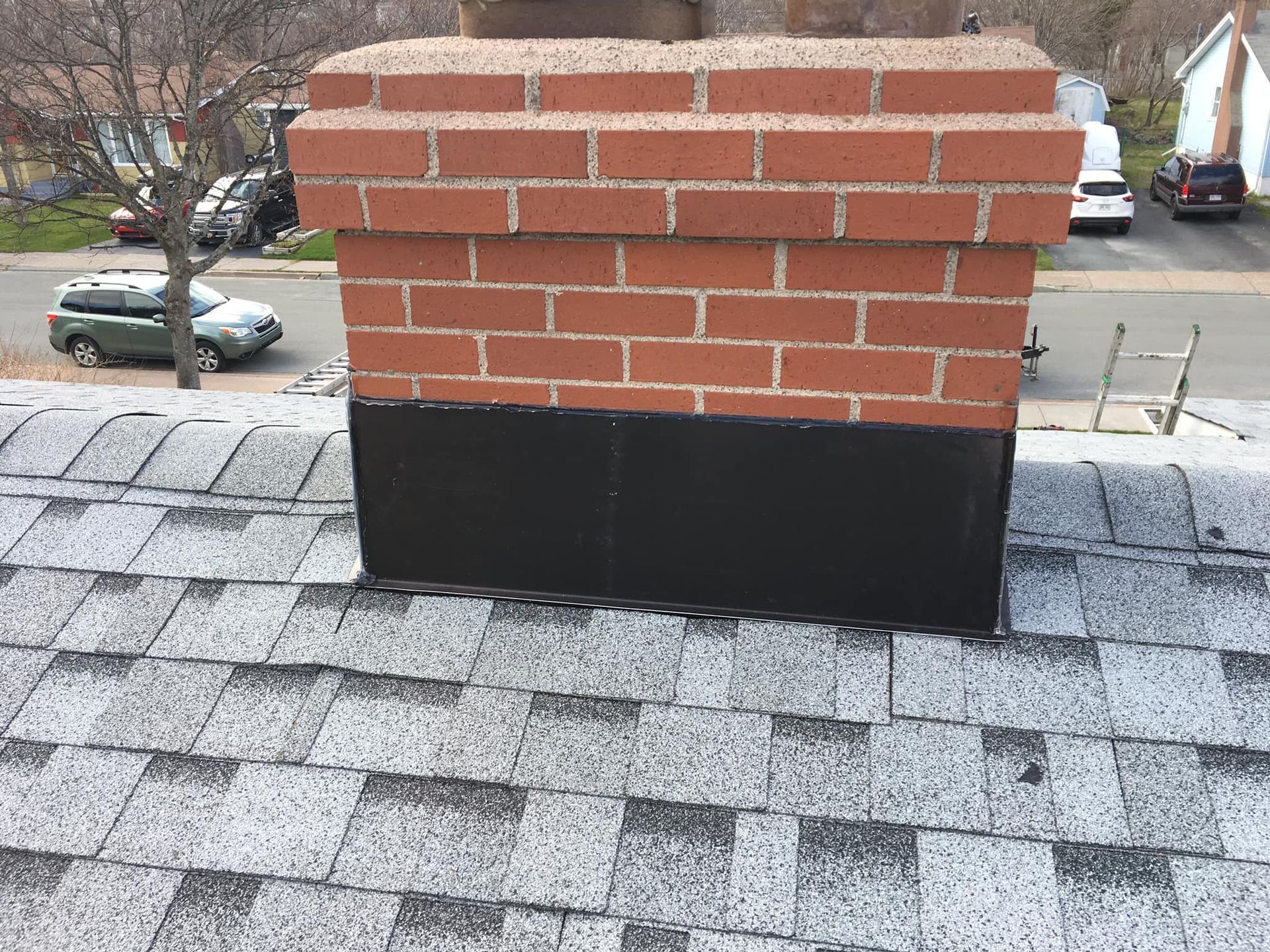 A brick chimney on a gray shingle roof, with a black painted base. Cars and houses are visible in the background.