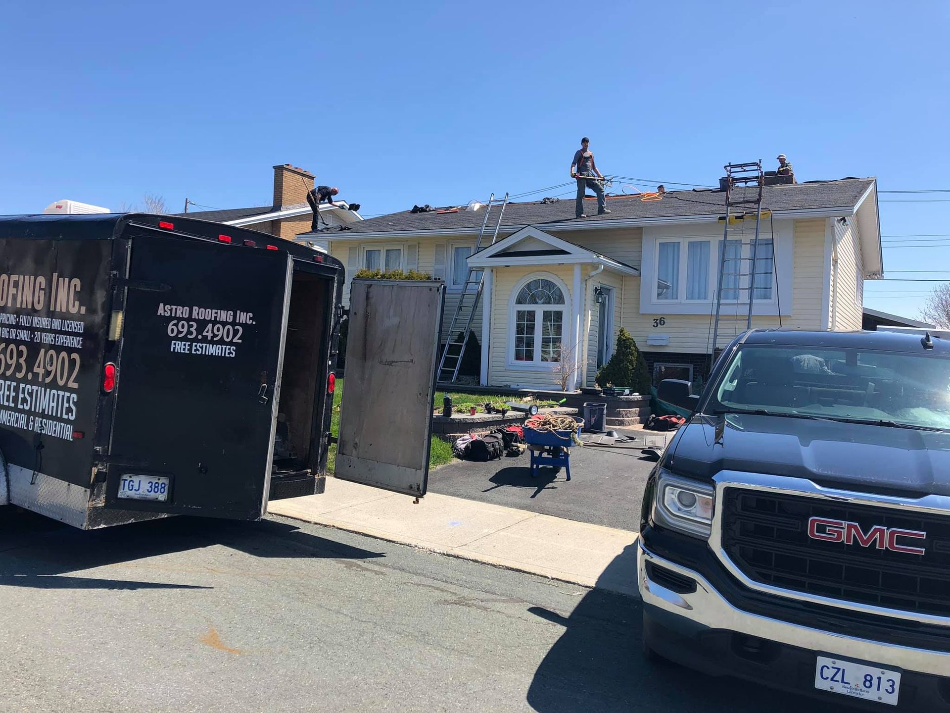 House with workers replacing roof, trailer, and GMC truck parked in driveway on a sunny day.