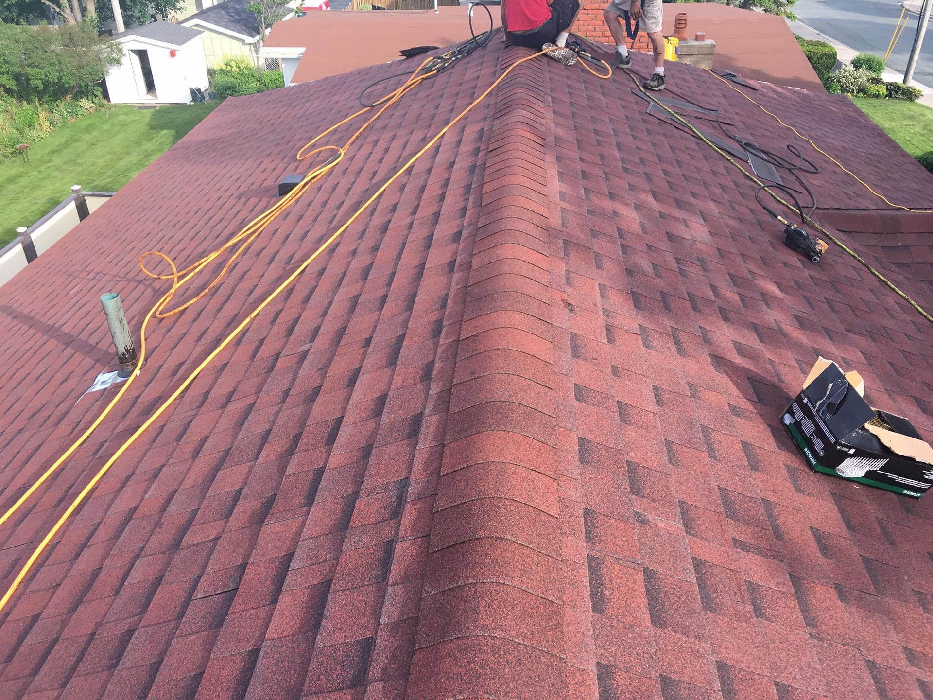 Workers on a reddish-brown shingled roof install cables. Green grass and buildings surround the home.