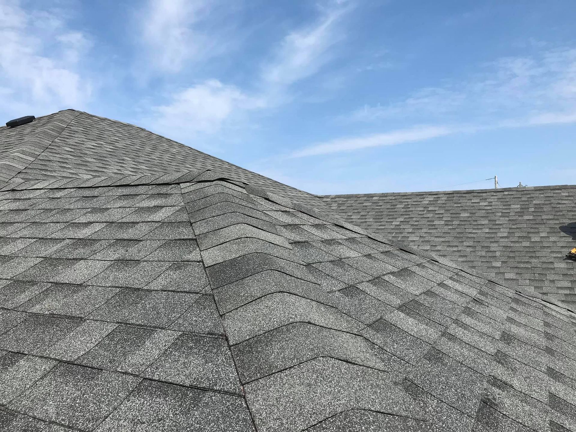 A close-up of an asphalt shingle roof, mostly gray, against a partly cloudy blue sky.