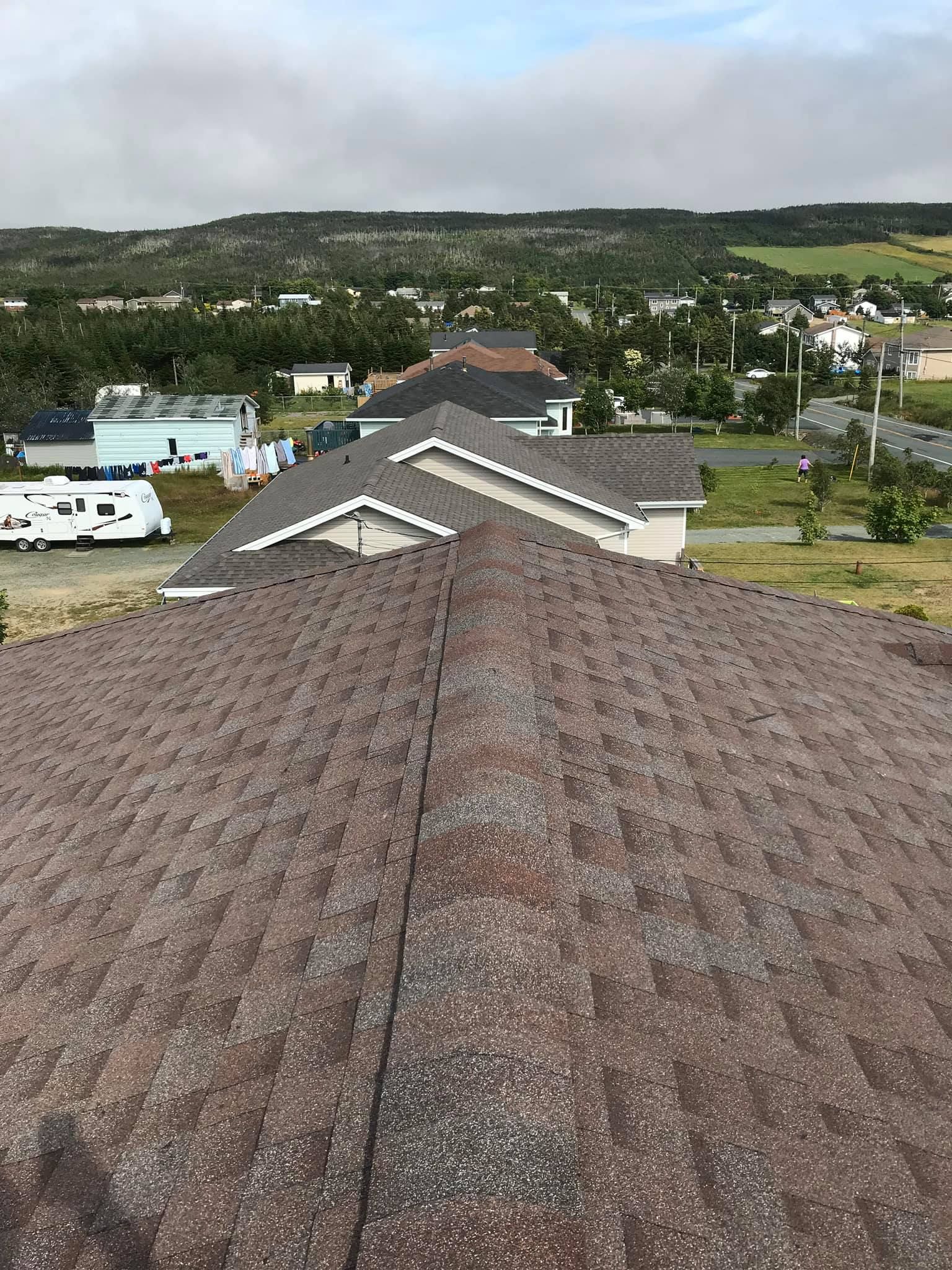 Brown shingled roof with a ridge, overlooking a residential area with houses and trees under a cloudy sky.