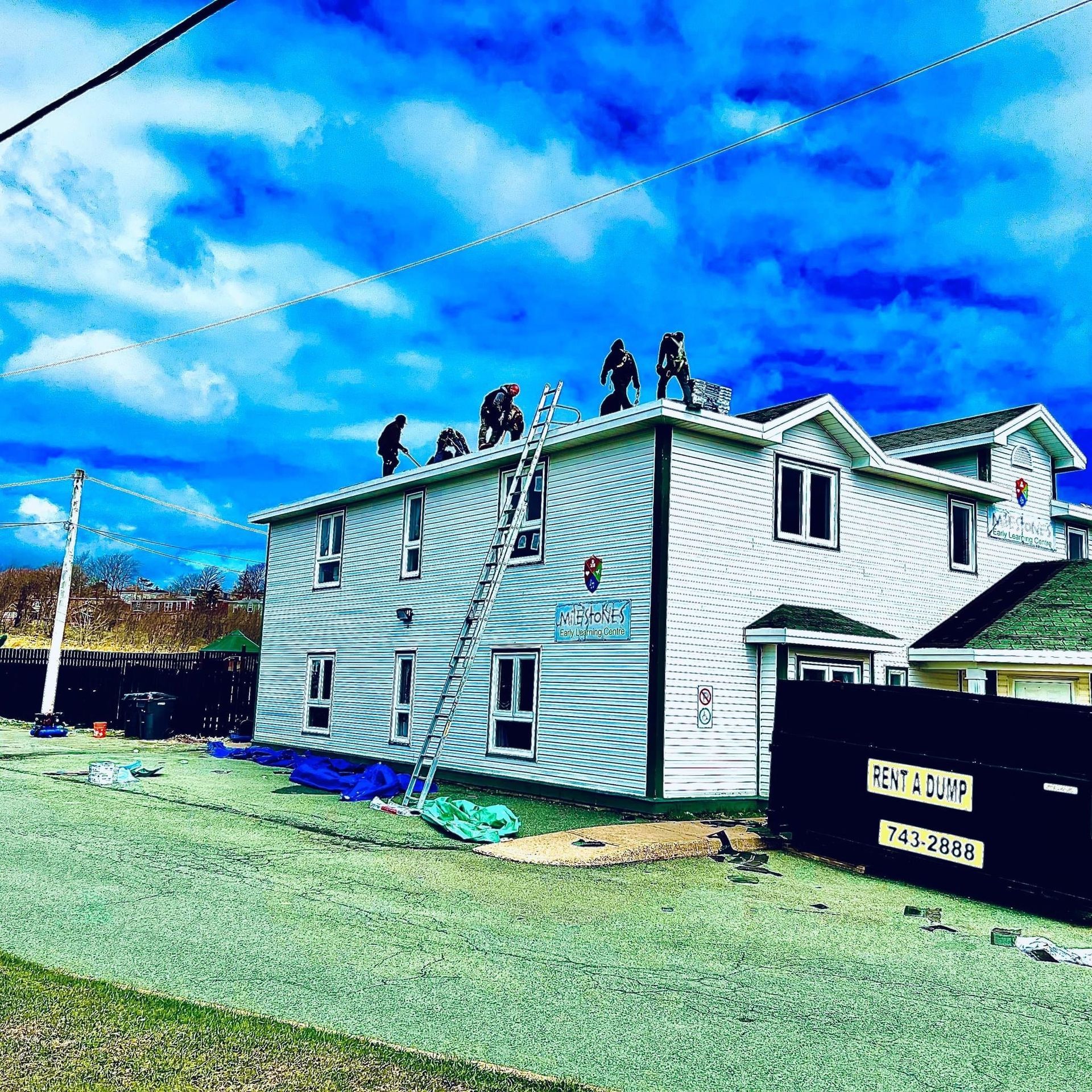 Workers on a white house roof under a bright blue sky with a ladder leaning against the side. A dumpster sits nearby.