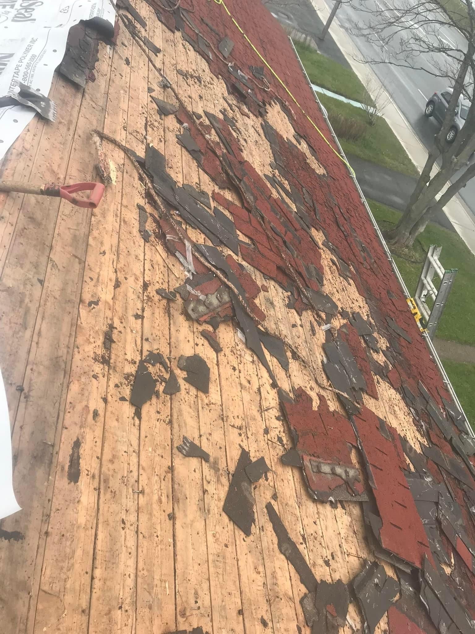 Damaged roof with missing shingles, exposing wood. Brown and red color scheme. A person is likely working on the roof.