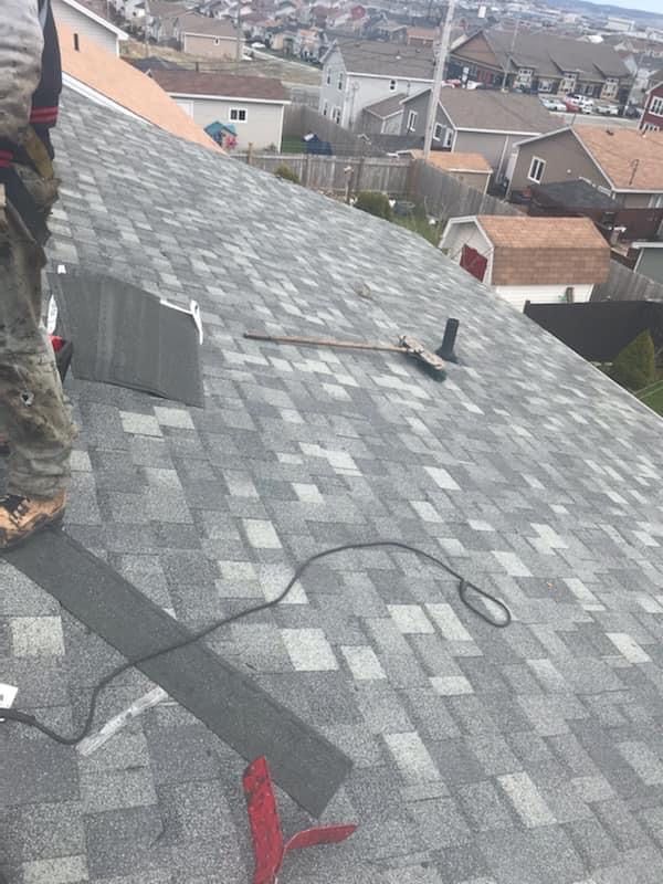 Roofer working on a gray shingle roof in a residential area.  Tools and safety equipment are visible.