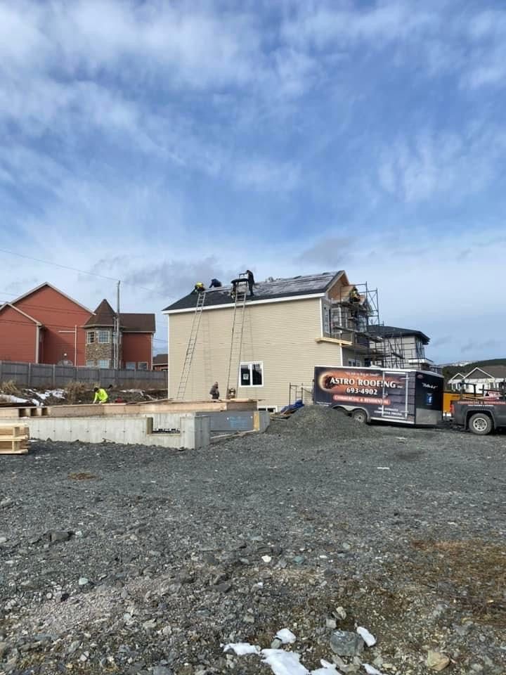 Construction site with a partially built two-story house. Workers are on the roof; other buildings and a trailer are nearby.