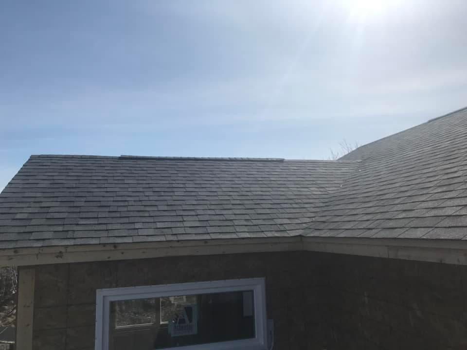 A roof with gray shingles on a sunny day. A white-framed window is below the roof line.