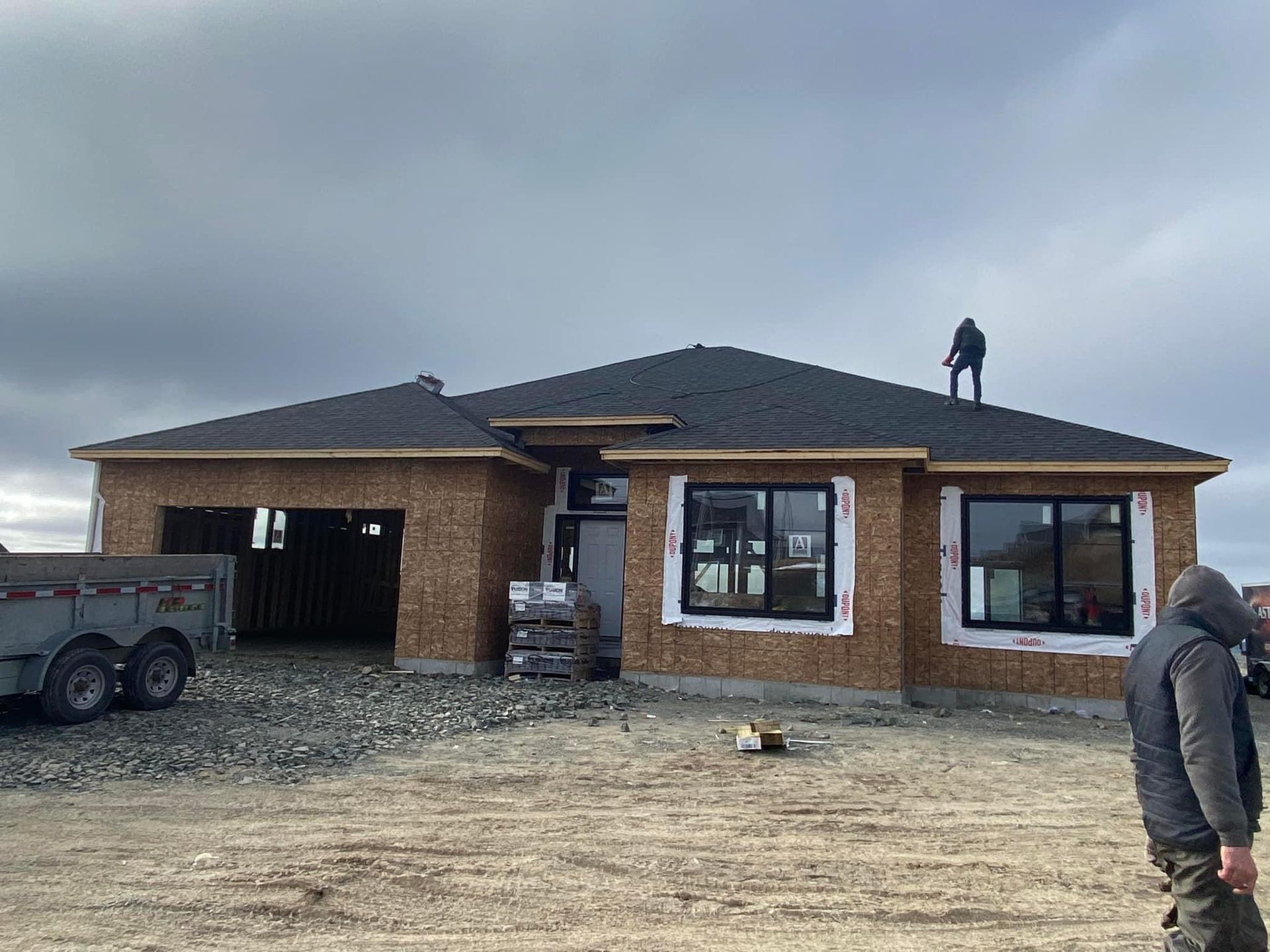 House under construction with dark shingles and windows, a worker on the roof, and another worker in the foreground.