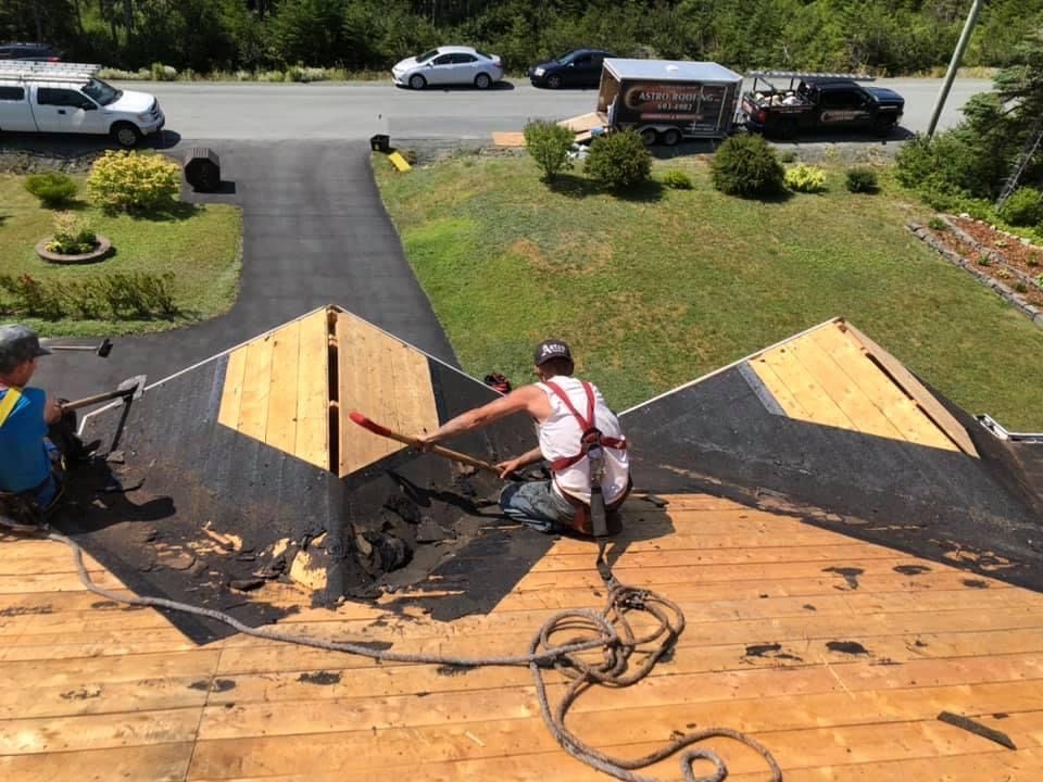 Two roofers working on a dark shingled roof, one kneeling, with tools. Cars and a yard are in the background.