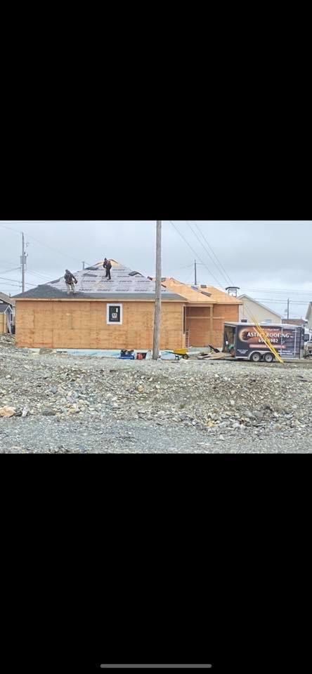 A house under construction on a gravel lot with workers on the roof. Gray sky overhead.
