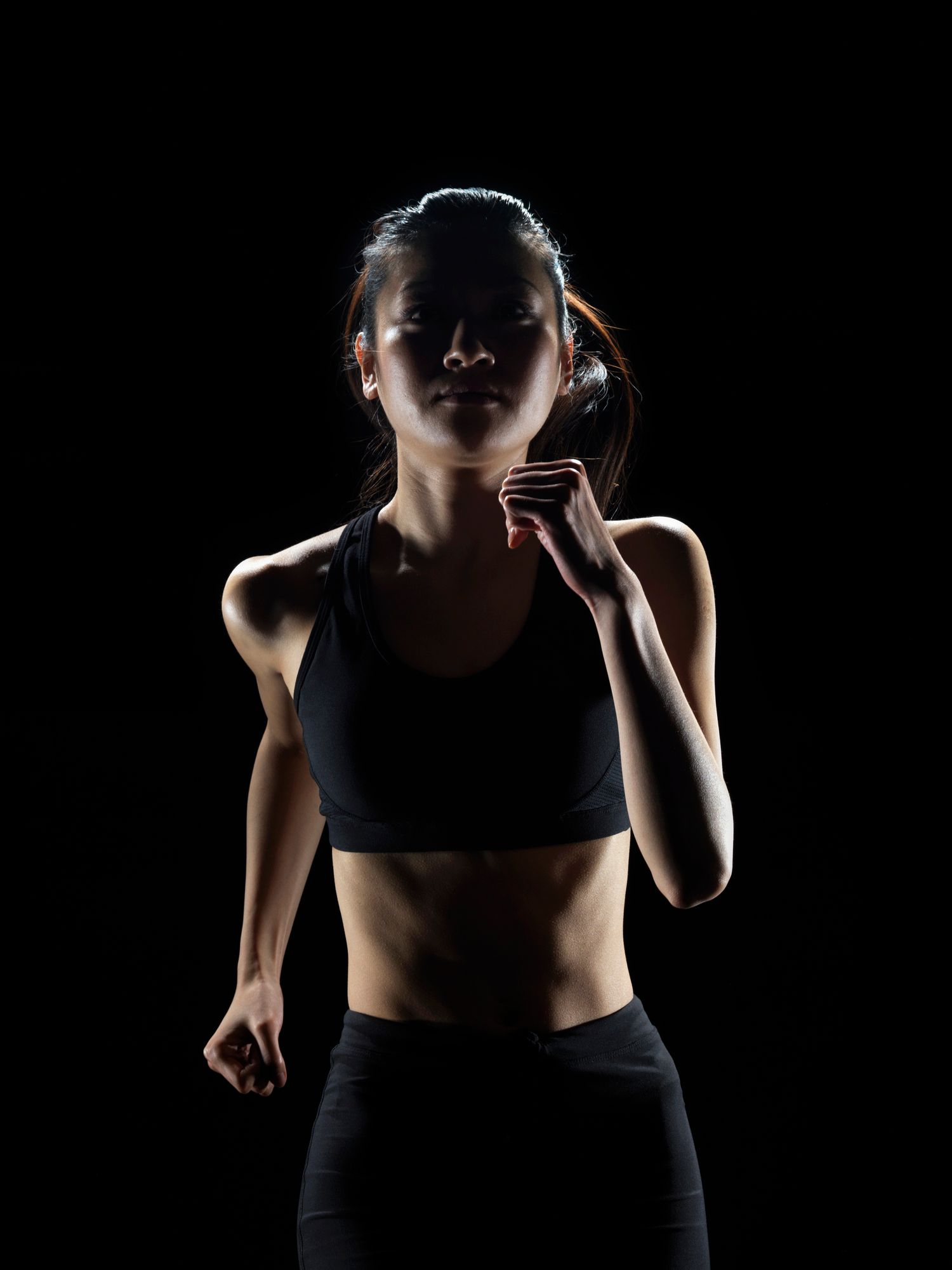 Woman in athletic wear running against a black background, lit from the front. Woman in athletic wear running against a black background, lit from the front.