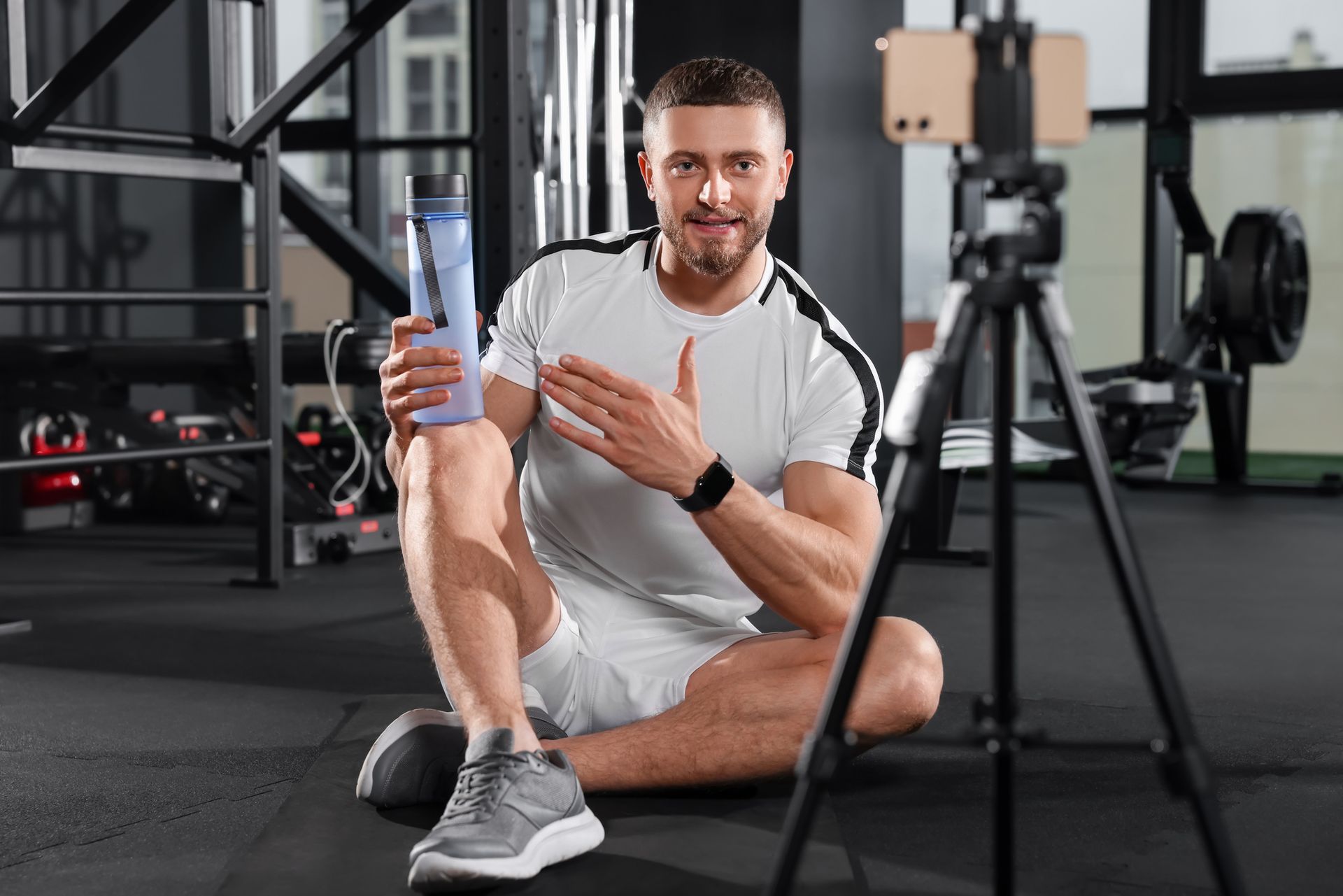 Man in gym with water bottle, gesturing towards phone on tripod. Black workout gear in background.