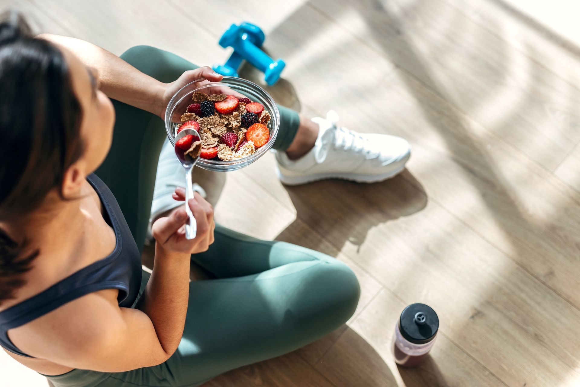 Woman in activewear eating a bowl of food on a wooden floor, with dumbbell and water bottle nearby.