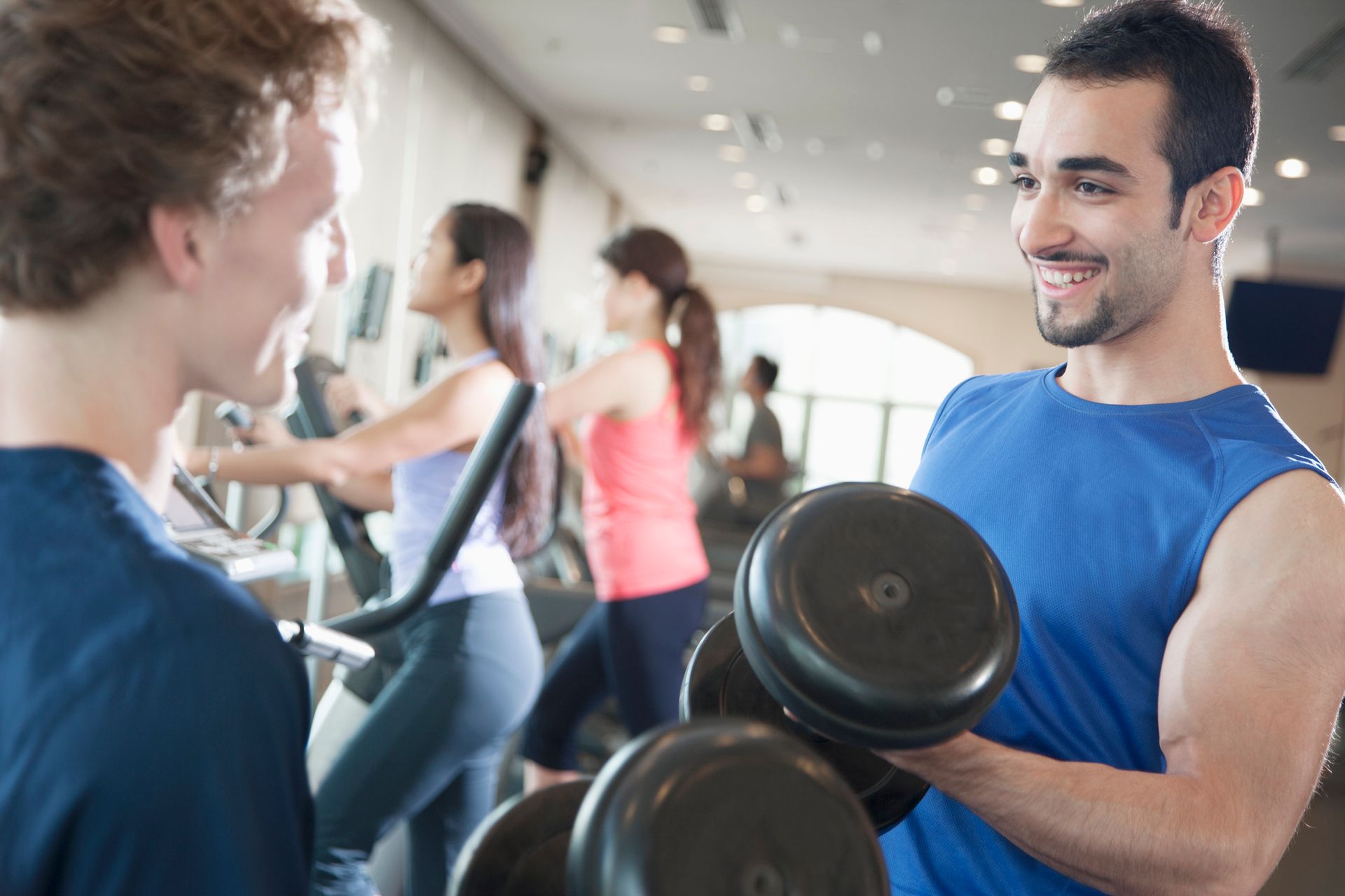 Man lifting dumbbells in gym, smiling at another man. People on exercise machines in background.