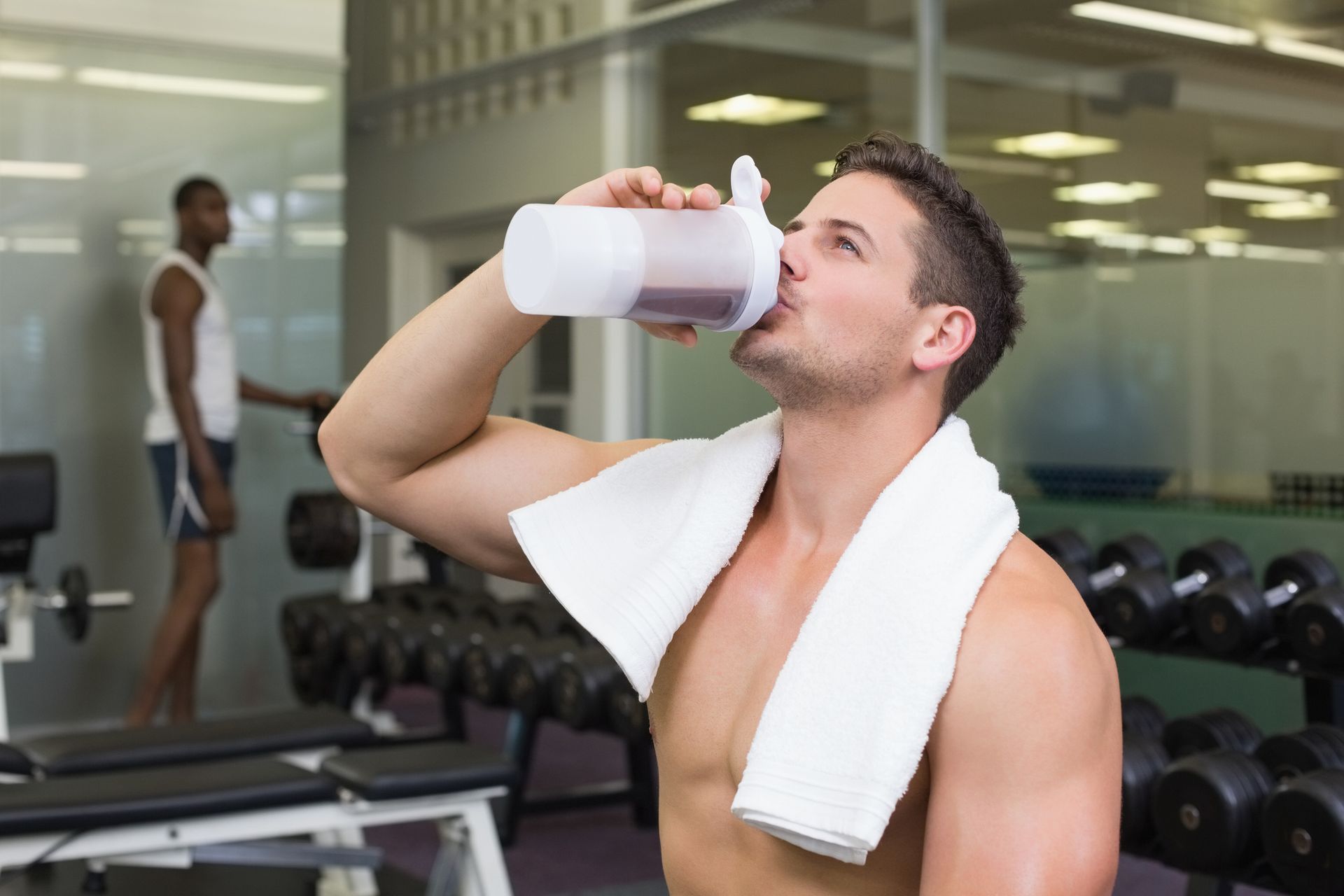 Man drinking from a protein shaker in a gym, white towel around his neck, another person in the background.