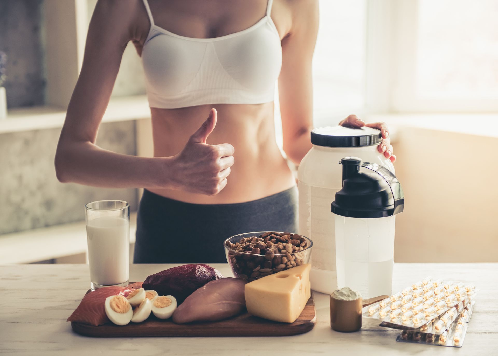 Woman with toned abs gives thumbs-up, surrounded by protein-rich foods, supplements, and shake in kitchen.