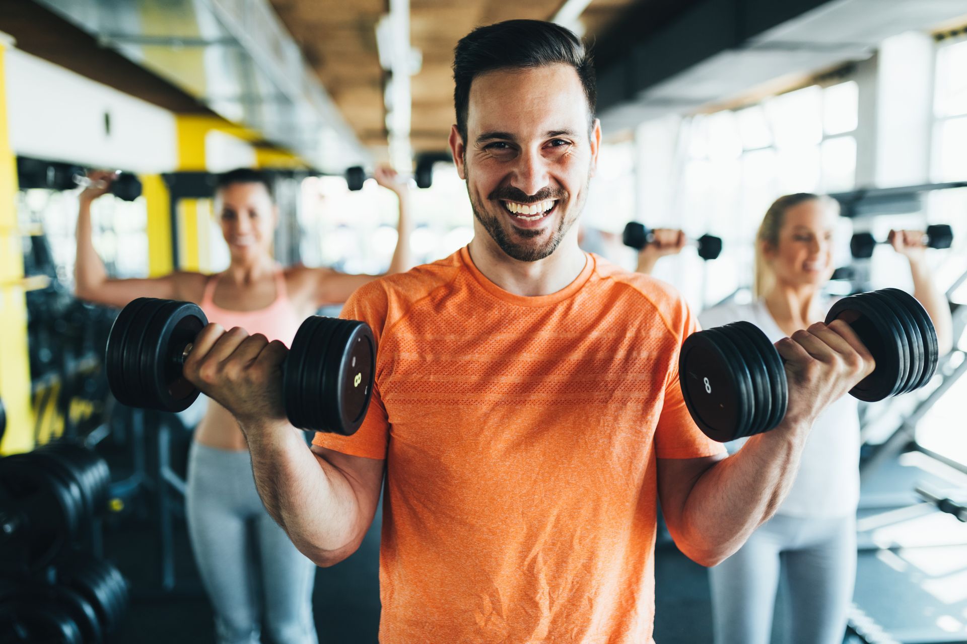 Man smiling, exercising with dumbbells in a gym. Two women in the background also lifting weights.