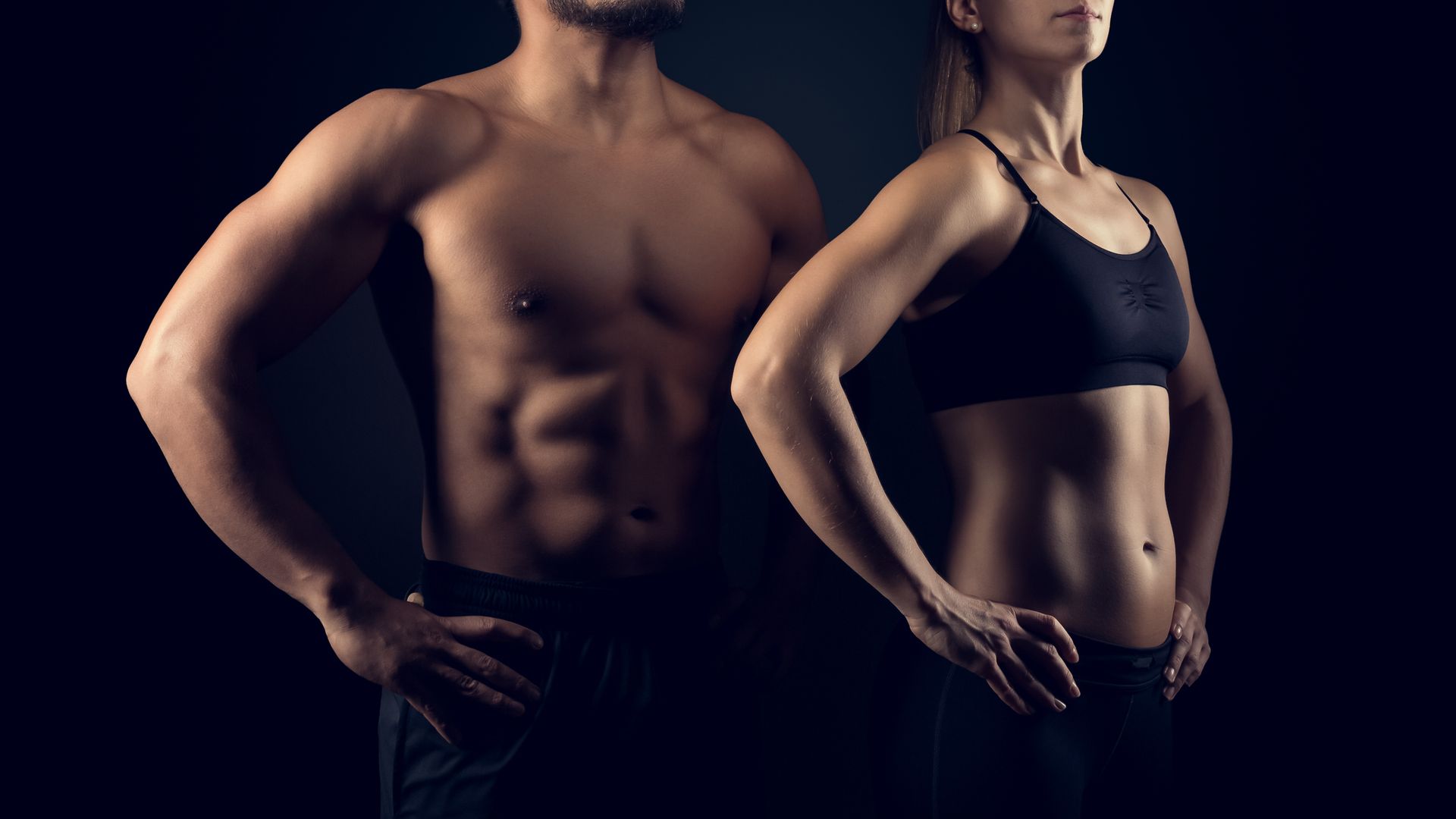 Man and woman with defined abs, hands on hips, dark background.
