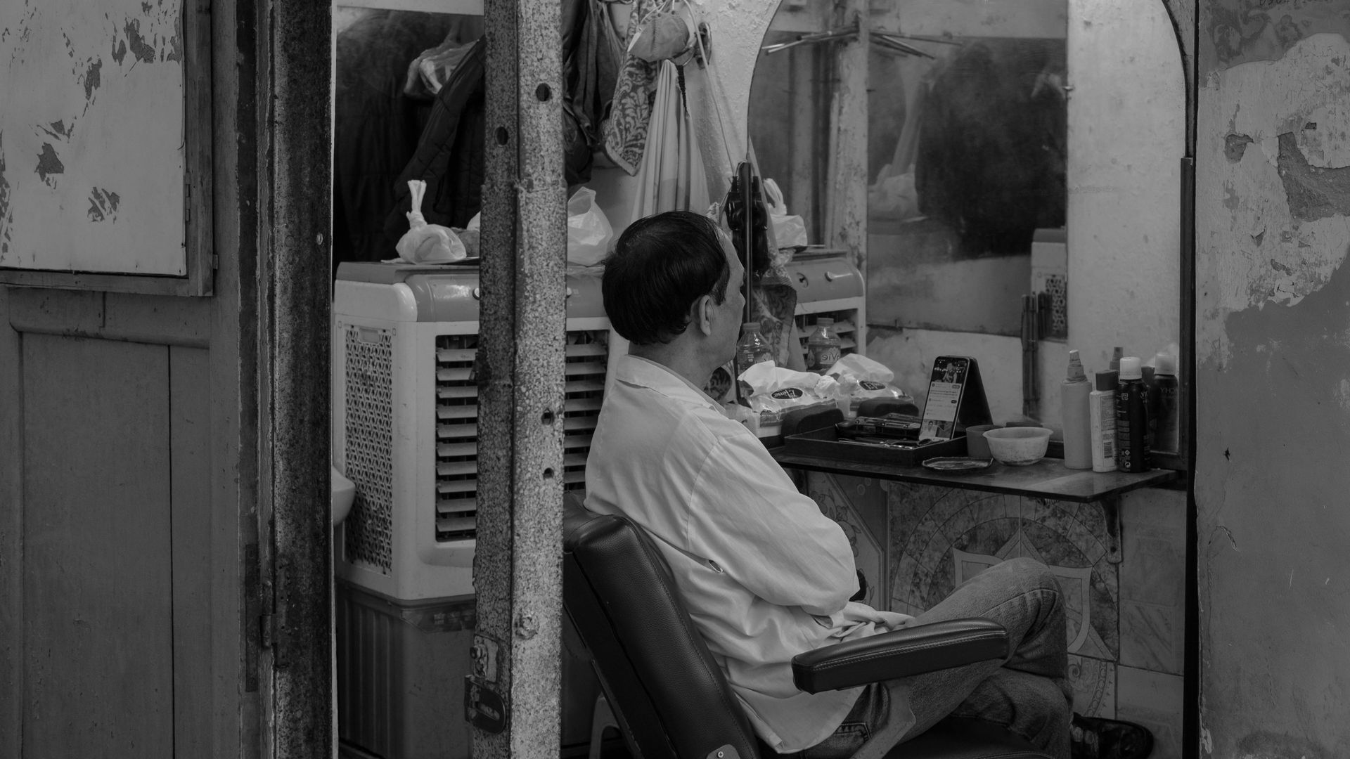 Barber in a dimly lit shop, seated with arms crossed, mirror behind him.