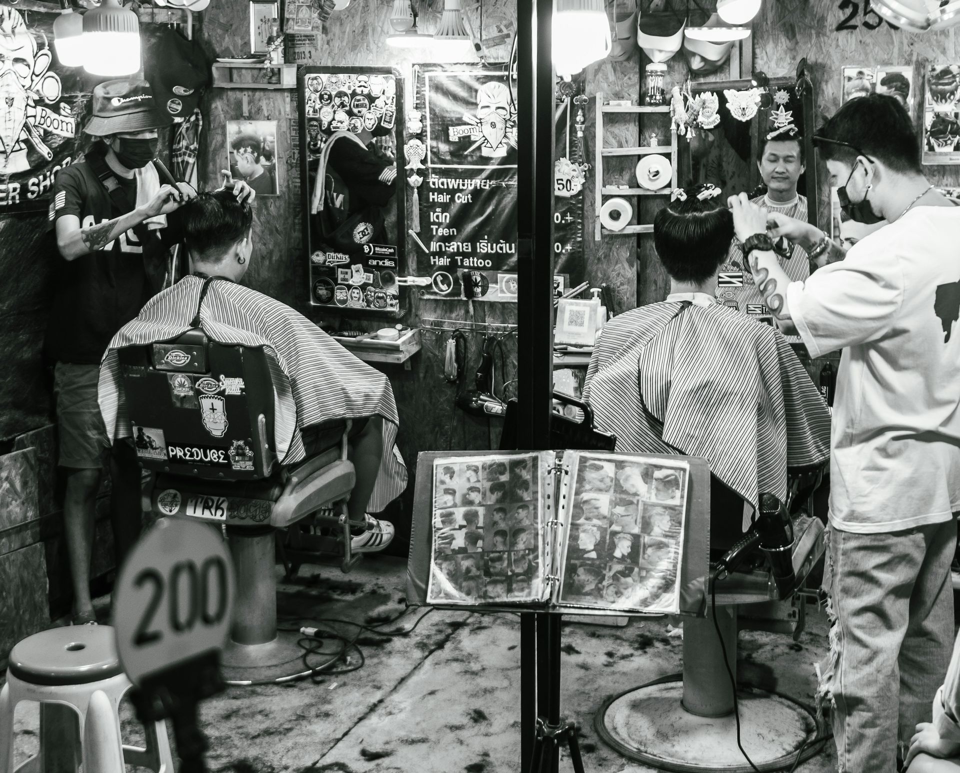 Black and white photo of a small barbershop. Two people get haircuts while barbers wear masks.
