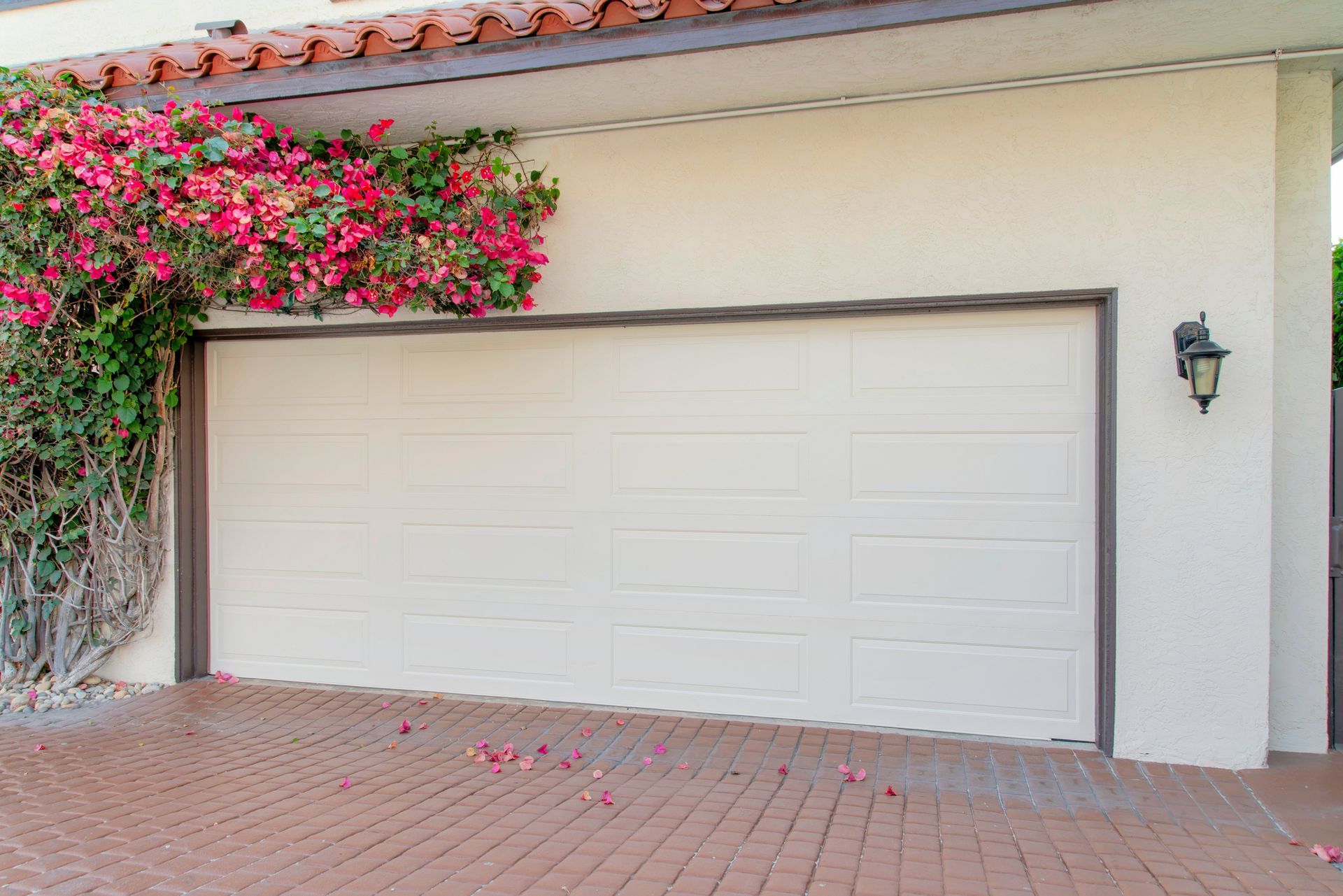 Porta del garage chiusa color bianco sporco con finiture marroni, ricoperta di fiori rosa rampicanti, appoggiata a un muro in stucco e a un vialetto in mattoni.