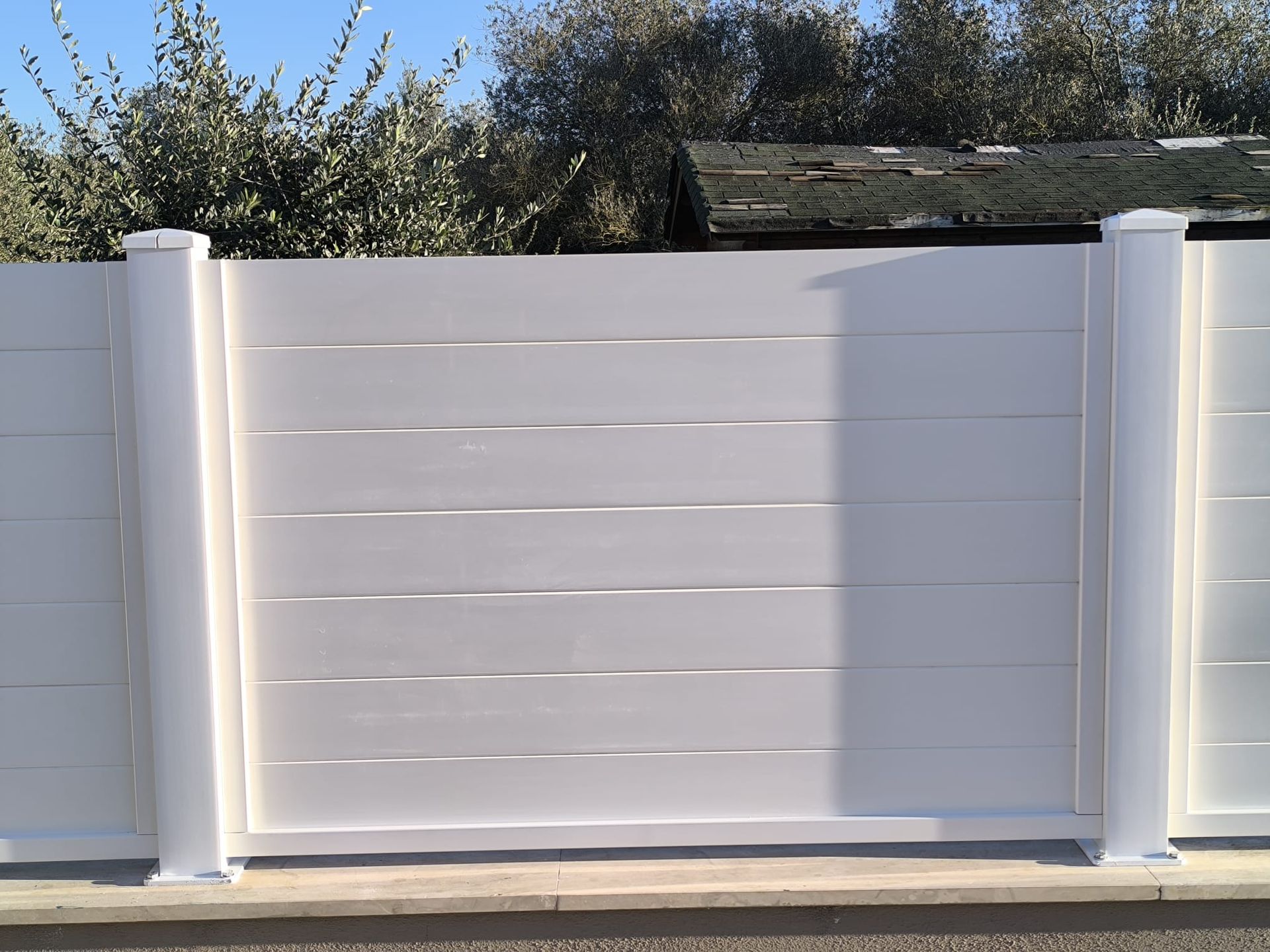 Recinzione bianca a doghe orizzontali con due pali di sostegno, posta su uno sfondo di fogliame sotto un cielo azzurro.