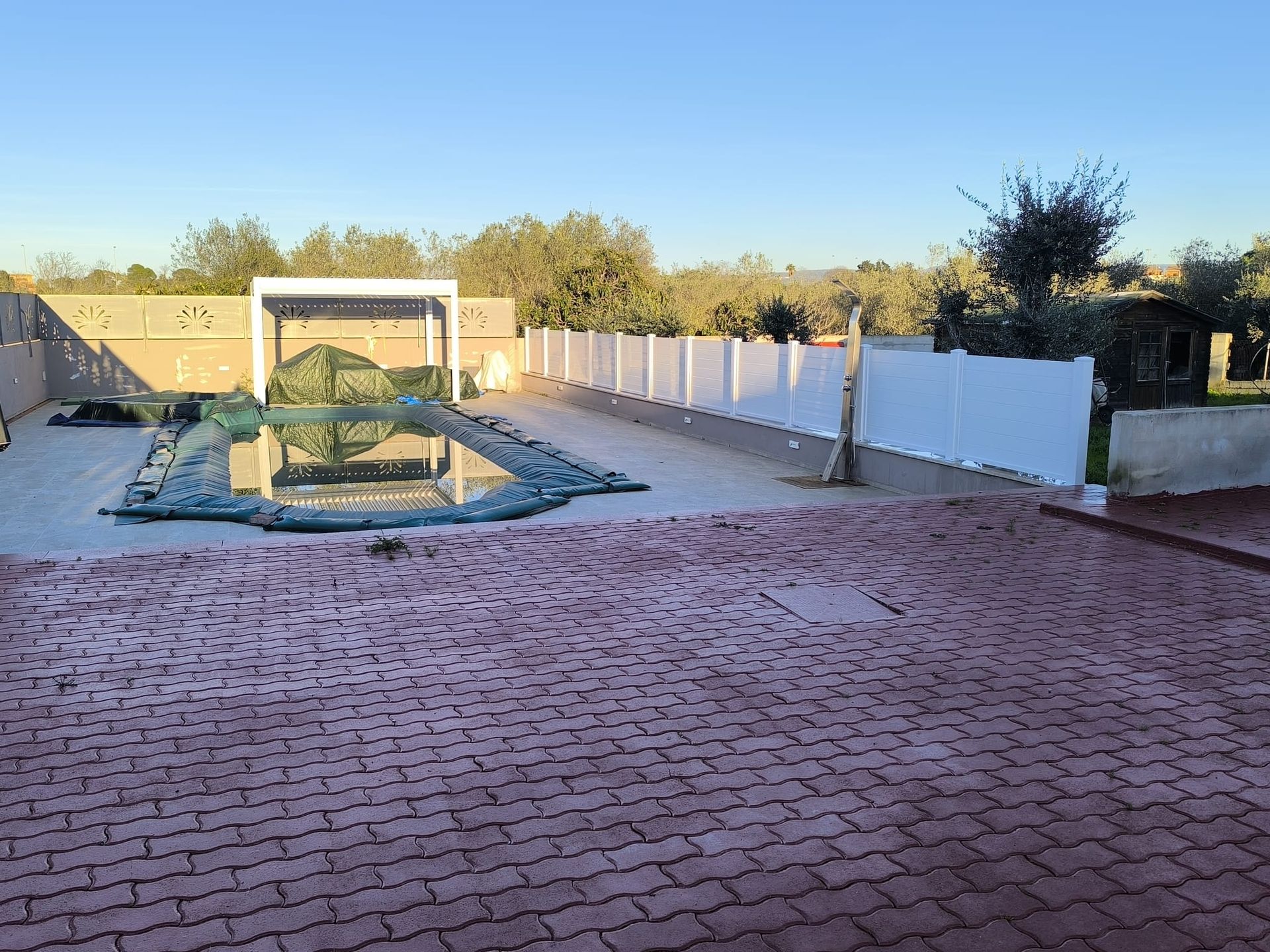 Area piscina con patio in mattoni, piscina coperta, recinzione bianca e pergolato.