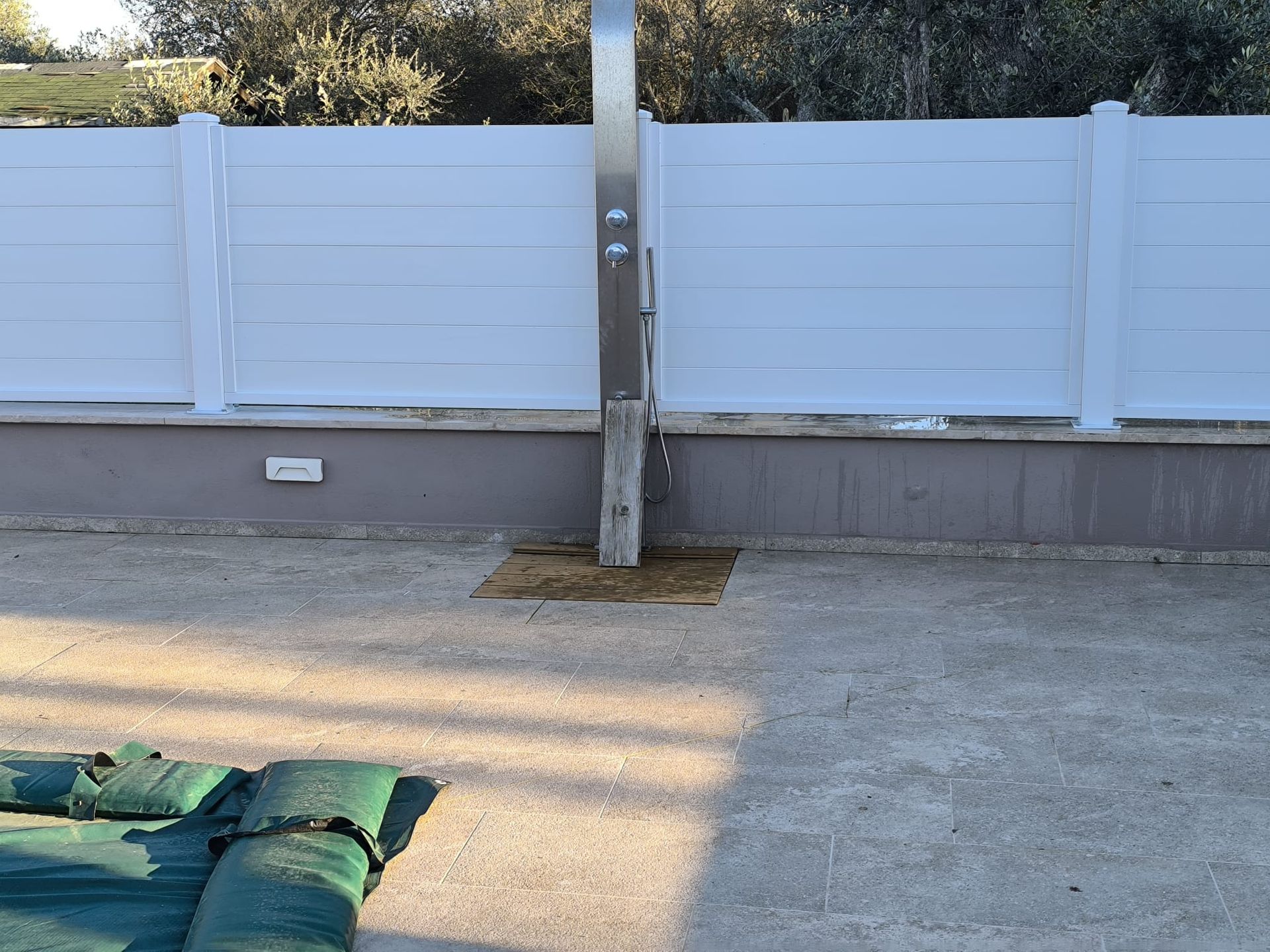 Doccia esterna contro una recinzione a doghe bianche. Patio in pietra grigia, tappeto verde in primo piano.