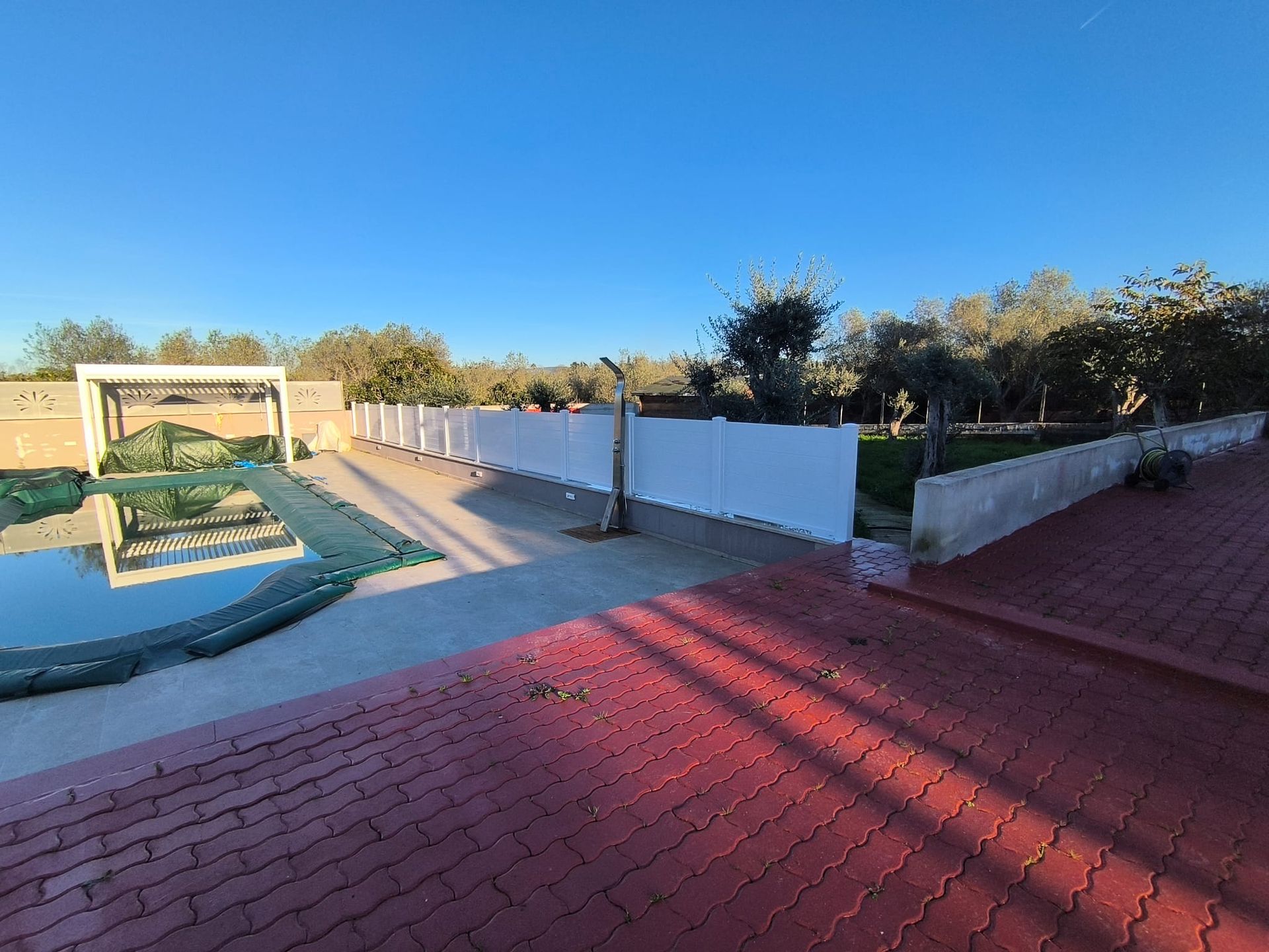 Scena all'aperto con una piscina, una recinzione bianca e un tetto di tegole rosse sotto un cielo azzurro e terso.