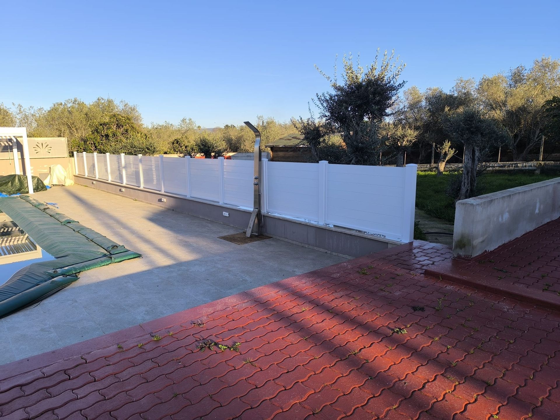 Recinzione bianca lungo un'area pavimentata vicino a una piscina e ad alcuni alberi. Patio in mattoni rossi in primo piano.